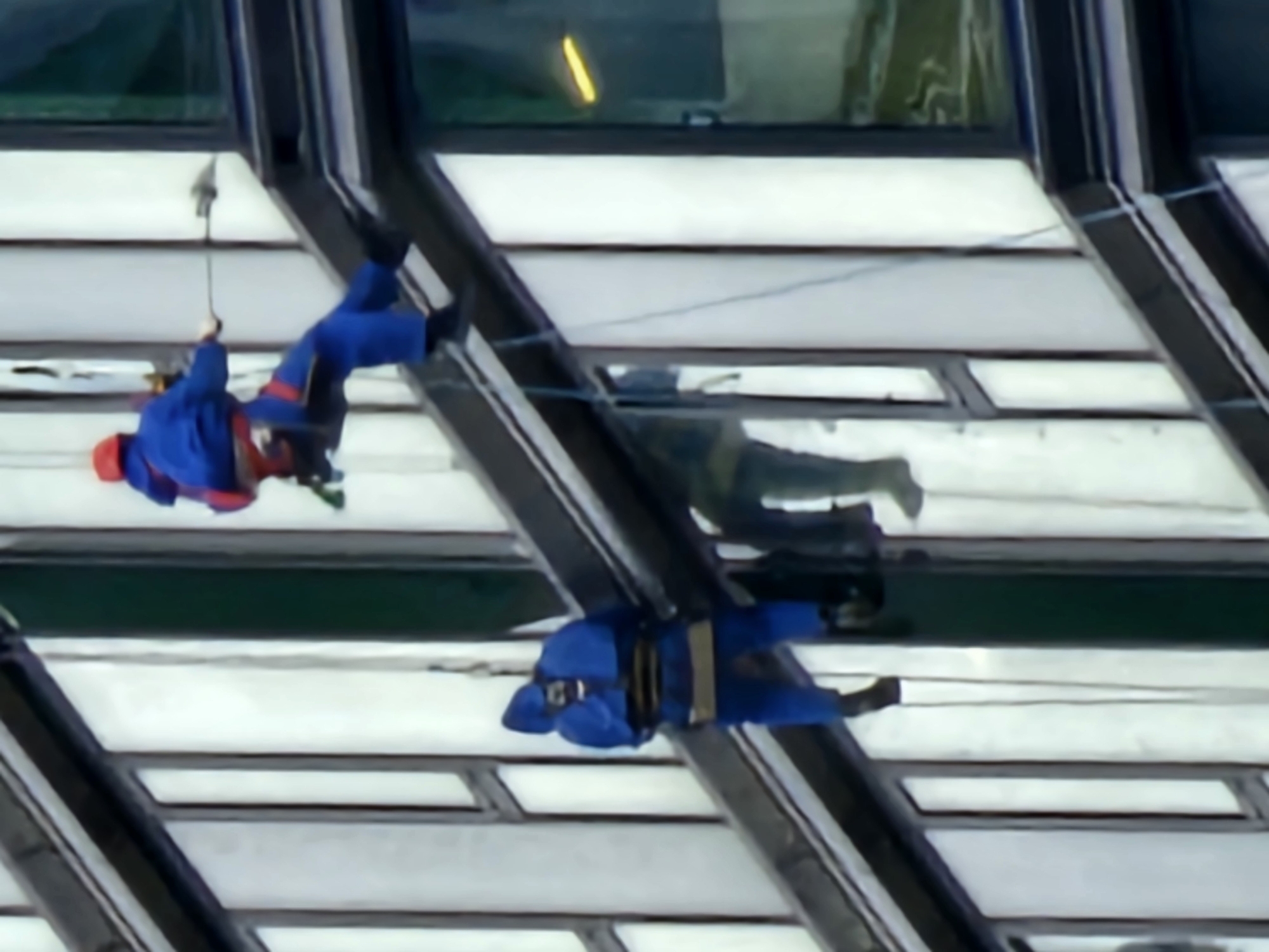 The image shows two individuals dressed in blue jumpsuits with red accents, rappelling down the side of a modern building. The building features large, reflective windows and dark structural elements. The first individual, higher up and further to the right, is wearing a red helmet and appears to be engaged in some sort of task, holding a cleaning implement or tool. The second individual, positioned below and to the left, is also wearing a hooded blue jumpsuit and is secured by ropes. The scene suggests window cleaning or maintenance work on a tall structure in an urban environment. The background is out of focus, indicating the image was taken from a distance or with a shallow depth of field. No specific Moscow landmarks are visible, but the architecture is consistent with contemporary city construction.