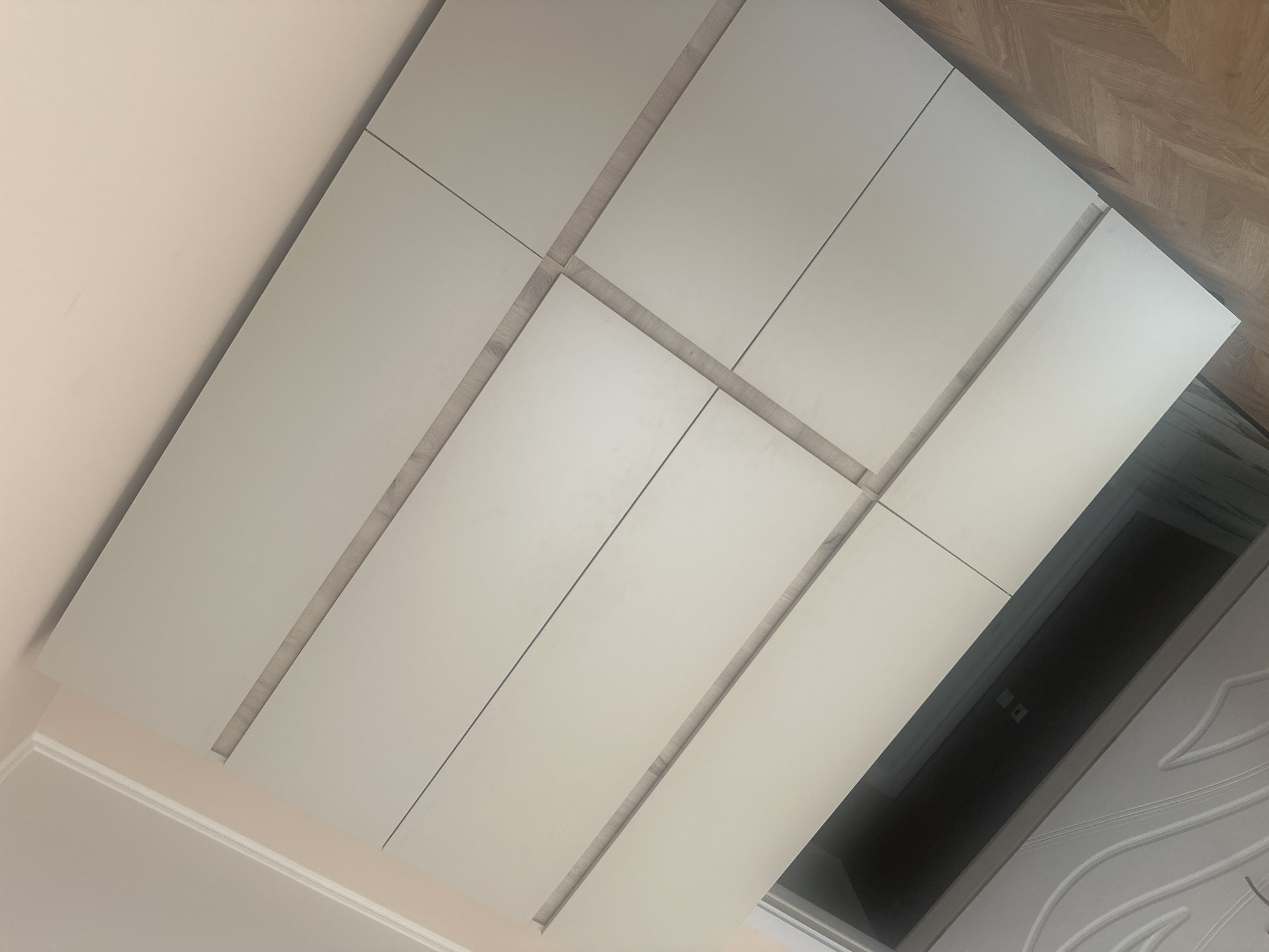 The image depicts a section of a room with a wall decorated with large, light-colored rectangular panels. These panels are arranged in a geometric pattern and have subtle grout lines between them. The wall appears to be part of a modern interior design. In the background, a doorway leads into a darker space, possibly a closet or another room, with electrical outlets visible on the wall within. The floor is a wooden parquet pattern. The lighting suggests an indoor environment. The image was taken in Plovdiv, Bulgaria.