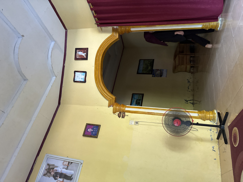 The image displays an interior residential scene in the city of Pante, Indonesia. The room features pale yellow walls with a dark red border along the white ceiling, which exhibits decorative molding. The floor is composed of light-colored square tiles, partially covered by a maroon rug with a lighter central pattern in the foreground.

On the left wall, a large silver-framed photograph depicts a man and a woman in formal attire. Below it, a smaller brown-framed portrait shows a man in a military-style uniform. A white electrical outlet with a plugged-in cord is visible below the smaller portrait.

A prominent arched doorway, decorated with yellow and white columns and trim, provides access to an adjacent room. Above the archway, two smaller framed pictures are mounted. A small decorative ornament hangs from the left column of the archway. To the right of the archway, a maroon curtain is partially drawn.

A black pedestal fan with a red central hub stands on the tiled floor in the main room, positioned near the left wall.

Through the archway, the adjacent room reveals slightly darker yellow walls. Two framed pictures, one depicting the Kaaba and another a landscape, are visible on its far wall, along with a hanging calendar. Two light-colored, possibly rattan, chairs are also present in the background of this room.

A person is partially visible on the right, emerging from behind the maroon curtain. This individual is wearing dark clothing and a dark head covering, and is barefoot, with their right lower leg and foot visible as they step into the main room.
