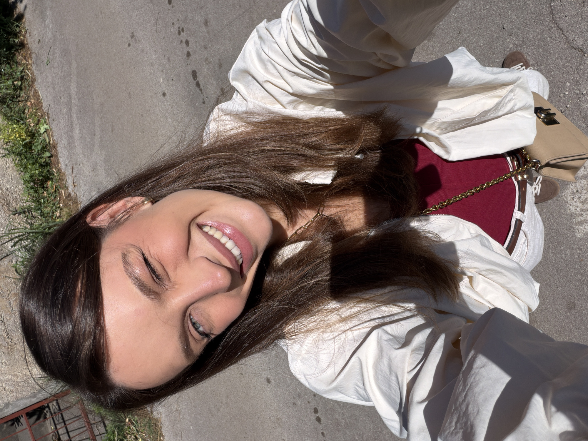 A woman is seen in a selfie taken at an upward angle. She is smiling and wearing a white jacket over a burgundy top. A beige purse with a gold chain is draped across her body. The background features a sidewalk, with hints of greenery and what looks like a low fence. Given the woman's appearance and the general environment, it is plausible that this image was taken in Budva, Montenegro.