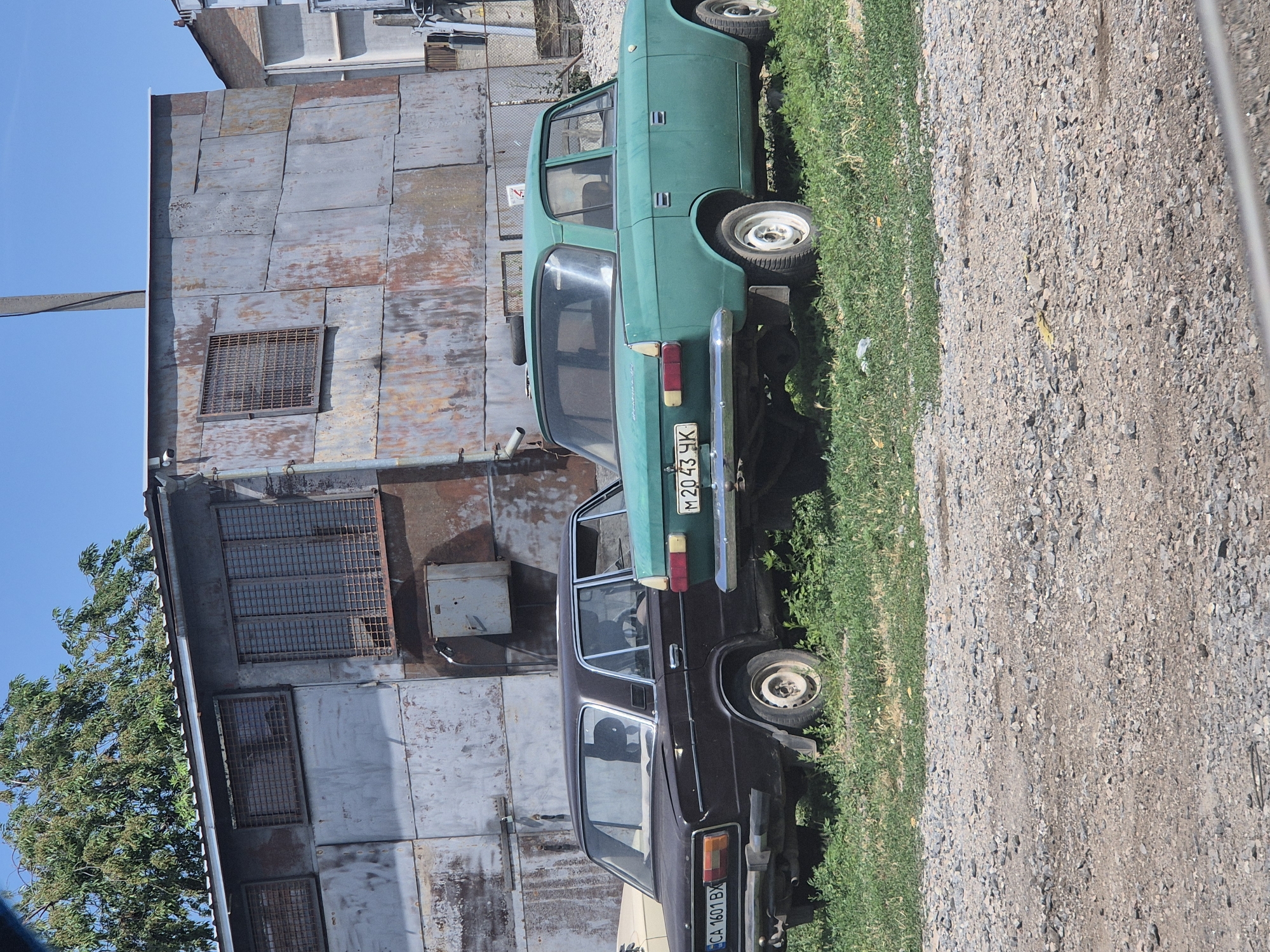 Two vintage cars, a dark brown sedan and a teal sedan, are parked on a grassy area next to a gravel road in front of a weathered metal building. The building appears to be a workshop or storage facility, with large, rusted metal panels and barred windows. The cars are old models, likely from the Soviet era, with visible signs of wear and tear. The dark brown car has a license plate that reads "CA 1601 BX". The teal car has a license plate that reads "M 2043 ЧК". The scene is bathed in sunlight under a clear blue sky. The environment suggests an industrial or semi-rural setting.