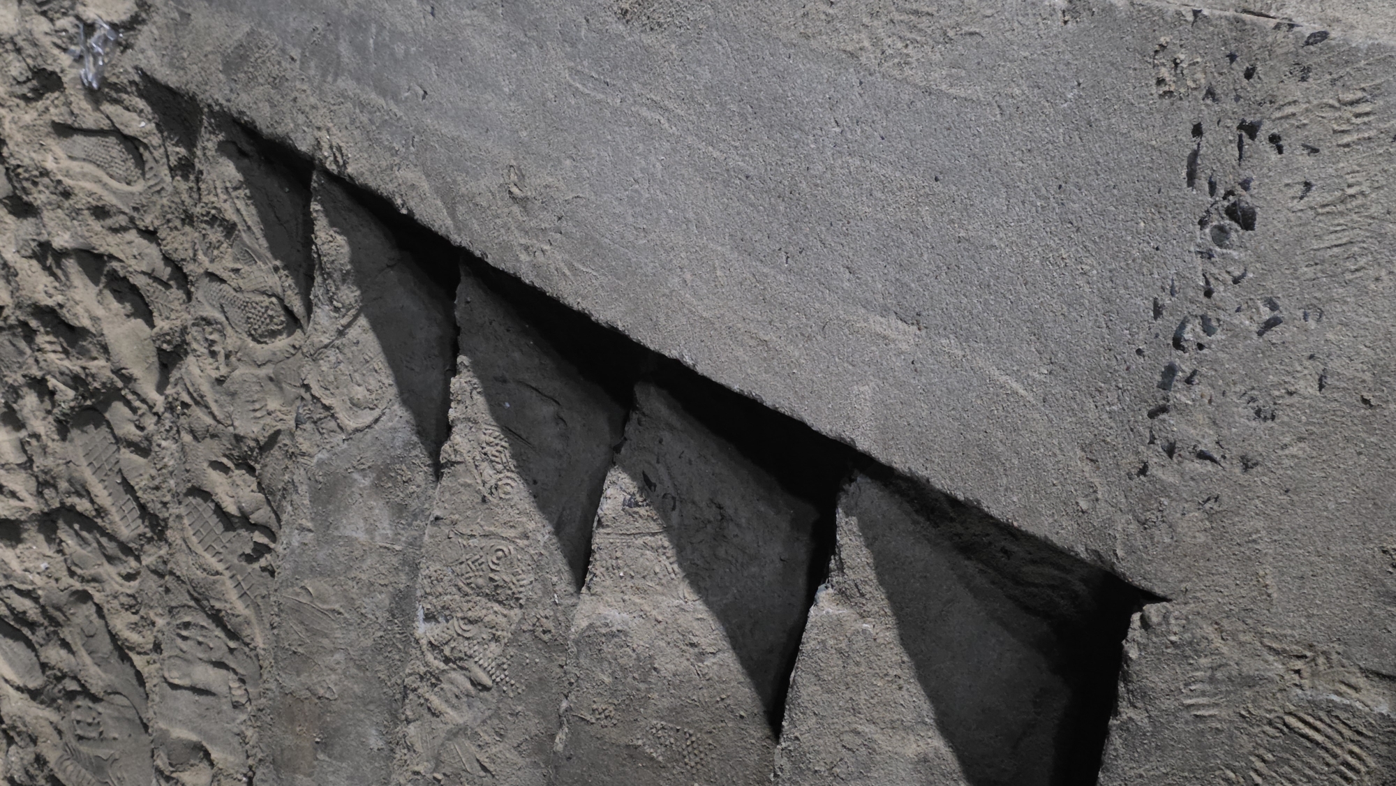The image presents an angled, close-up view of a rugged, grey-toned environment, possibly in Nha Trang, Vietnam. A large, flat, concrete or stone slab occupies the upper and right sections, displaying a coarse, gritty texture with minor surface variations and scattered indentations, possibly small stone chips or debris marks.
Below this main slab, a series of sharp, dark, triangular voids are visible, separating several irregularly shaped, pointed forms that jut out from the lower ground. This lower surface, especially on the left and between the pointed protrusions, consists of a light grey, dusty or sandy material. It is heavily marked with numerous shoe prints of varying sizes and patterns, alongside some discernible tire tracks, indicating frequent human and vehicular activity. The patterns on the shoe soles are diverse, ranging from geometric grids to more intricate designs. Some of the pointed earth structures also show clear shoe imprints. A small amount of white, fibrous debris is visible on the far left.
The scene is brightly lit by direct sunlight, casting strong, well-defined shadows beneath the upper slab and behind the pointed forms, suggesting a clear, sunny day. No people or animals are present, but their passage is evident through the extensive imprints. No visible text is present in the media.