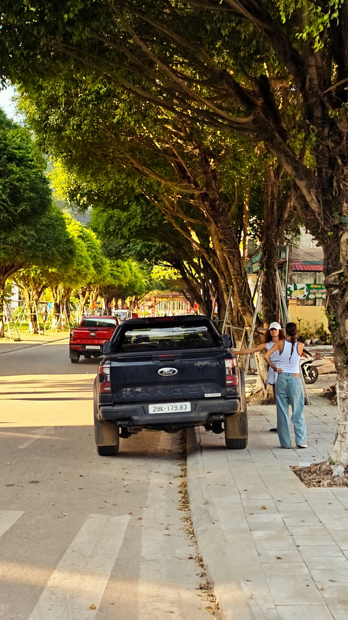 The image shows a street scene in Lao Cai Ward, Vietnam. A dark blue Ford Ranger pickup truck is parked on the side of the road, partially obstructing a pedestrian crosswalk. The truck's license plate reads "29K-179.83." Two women stand on the sidewalk beside the truck. One woman wears a white top, blue jeans, and a white baseball cap. The other woman wears a white top and has her hair in braids. In the background, a red pickup truck travels down the street, which is lined with trees that form a canopy overhead. A motorcycle is parked on the sidewalk beyond the women. A building with a sign is visible further down the street. The lighting suggests it is late afternoon.