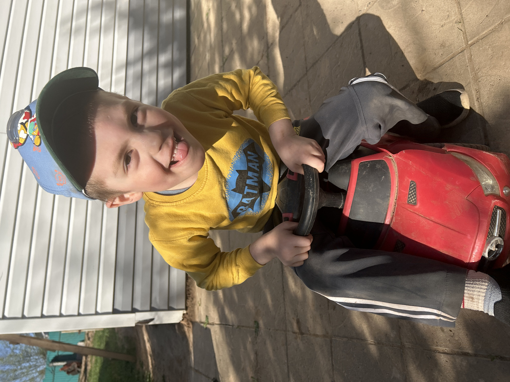 A young boy wearing a blue baseball cap, a yellow sweater with a Batman logo, and grey sweatpants with white stripes is sitting on a red toy car outdoors. He is sticking his tongue out and looking at the camera. The setting appears to be a residential area, with a white fence visible in the background and a tiled or paved surface beneath. Shadow patterns suggest sunny conditions. The location cannot be confirmed as being in the city of Фоки, Russia from the provided image.