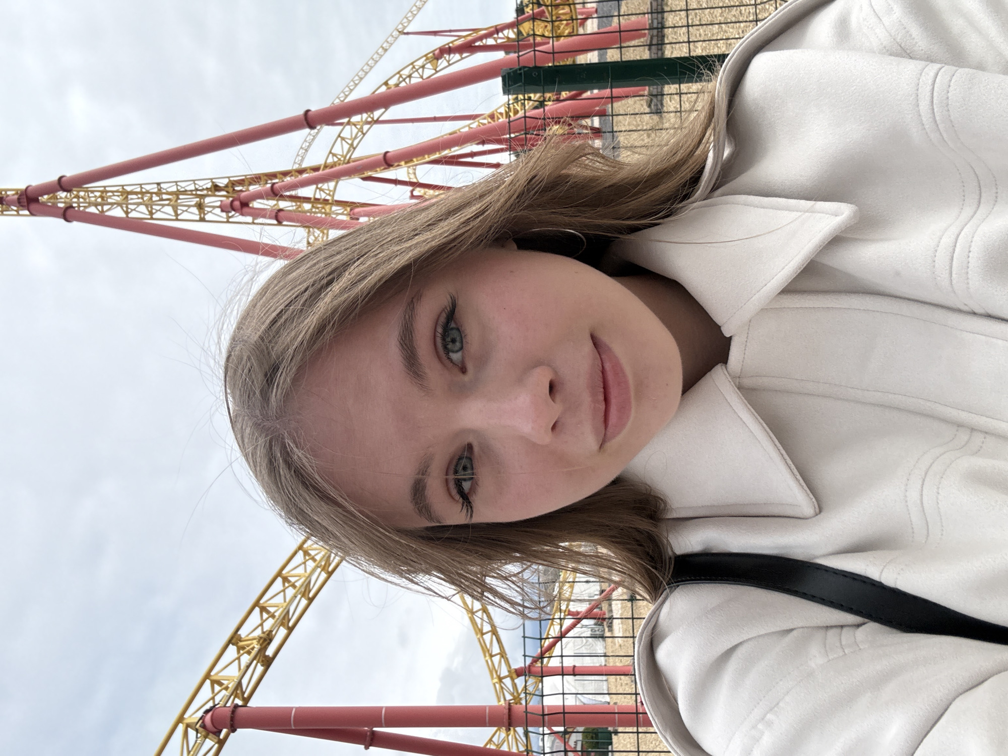 A young Caucasian female, approximately 18-25 years old, with light brown hair and blue-green eyes, is positioned centrally in a close-up selfie-style portrait. She wears a light beige or off-white collared jacket and small silver hoop earrings, looking directly at the camera with a subtle, closed-mouth smile. The background prominently features the "Quantum Leap" roller coaster, located at Sochi Park in the Adler District of Sochi, Russia. The roller coaster is characterized by robust red support structures and yellow truss-style tracks that ascend and curve against an overcast, light grey sky. A dark green metal fence with square mesh openings separates the foreground from the amusement park elements. Beyond the fence, dry, light-colored ground and a few white tent-like structures are partially visible.