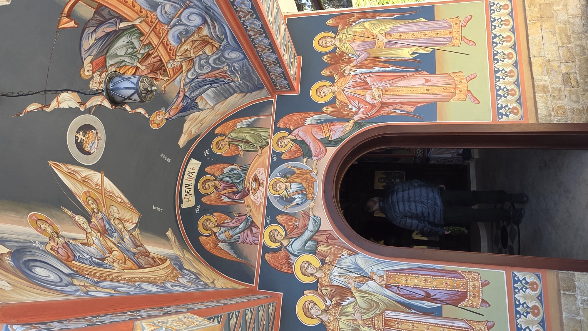 The image shows the interior of a church in Elenovo, Bulgaria. The walls and ceiling are covered in colorful Orthodox Christian iconography, depicting religious figures and scenes. A man stands in the doorway. He is wearing a blue jacket and dark pants. The doorway opens into the church's interior.