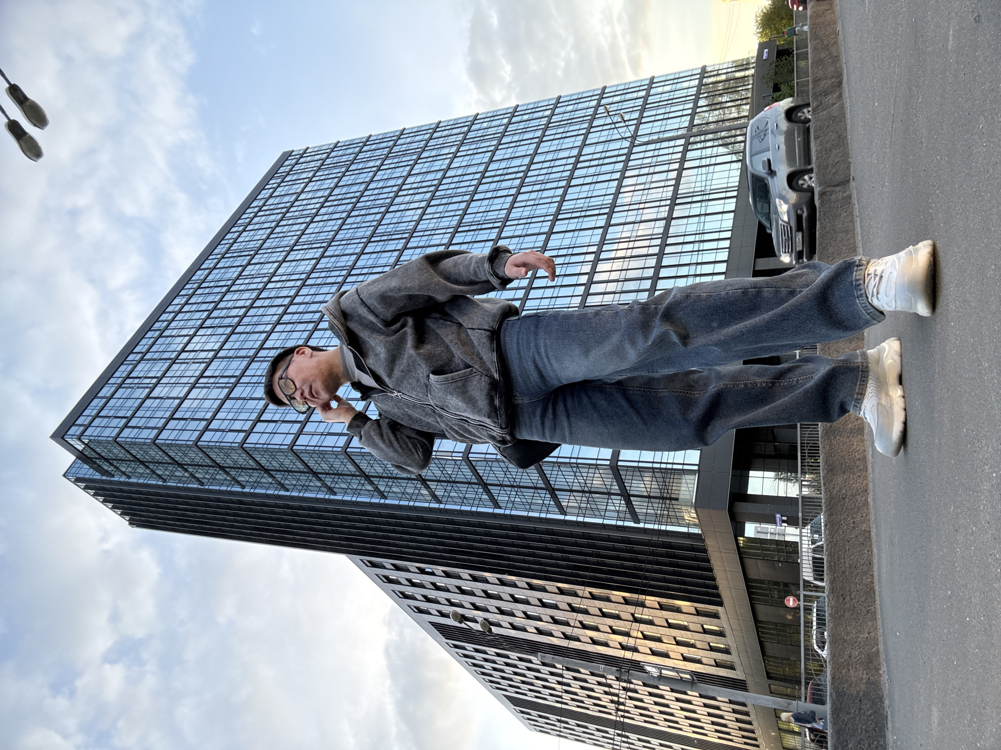 The photograph, originally rotated 90 degrees clockwise, captures a low-angle perspective of a person standing outdoors in an urban setting. The person, appearing male and of East Asian descent, is situated on what seems to be a sidewalk or street, wearing eyeglasses, a grey zip-up hoodie, blue jeans, and white sneakers. His right hand is raised near his ear, suggesting he is either talking on a phone or listening to something.
Dominating the background and left side of the frame is a striking modern high-rise building, characterized by its extensive glass facade and dark framing, reflecting the partly cloudy sky. Further in the distance, another building with a different architectural style, featuring numerous smaller windows, is partially visible.
The scene unfolds during the daytime, likely late afternoon or early evening, given the soft, warm light on some surfaces and the bright sky. A paved street or road runs parallel to the sidewalk, with a concrete curb separating the two. A dark vehicle is parked or driving on the street. In one of the cropped views, a red circular 'no entry' sign with a white horizontal bar is discernible near the street. Two streetlights are also visible against the sky in the upper left. The image conveys a moment of urban life amidst contemporary architecture.