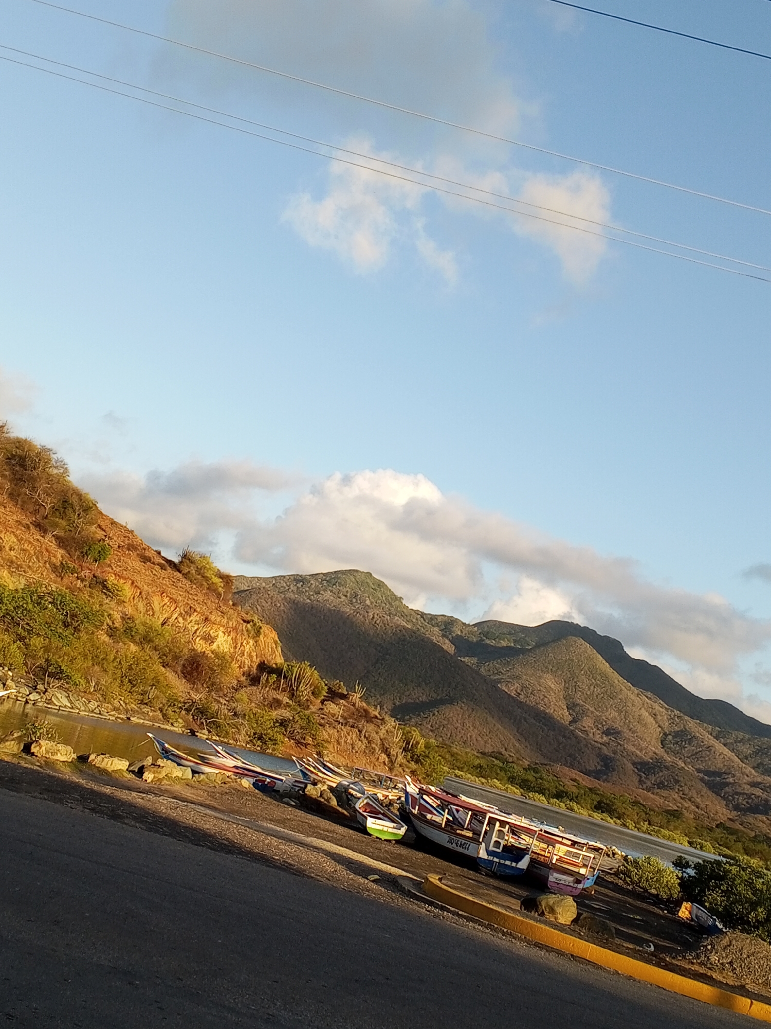 The image shows a roadside scene in Pedro González, Venezuela. In the foreground is a section of asphalt road with a yellow painted curb. Beyond the curb lies a shoreline with a collection of small, colorful fishing boats resting on the gravelly shore. Some of the boats are adorned with canopies and painted details. Behind the boats is a body of water, likely a bay or inlet, bordered by vegetation. Rising in the background are dry, mountainous hills under a partly cloudy, light blue sky. Overhead, power lines stretch across the sky. The time of day appears to be late afternoon, with warm light casting shadows on the landscape.