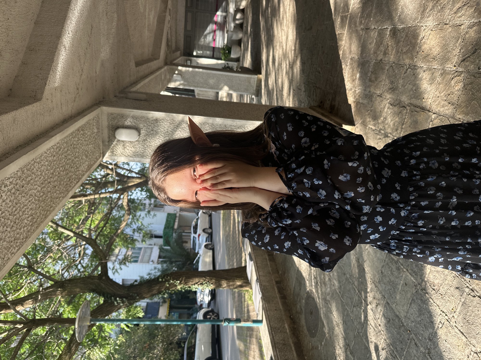The image is an outdoor shot of a person covering their face with their hands, positioned within a covered walkway. The person appears to be wearing a dark-colored dress with a light-colored floral pattern and pointed ears, giving them an elvish appearance. The walkway has a concrete structure, with a series of supporting columns and horizontal beams, casting shadows on the stone-paved ground. Sunlight filters through the trees and building structures, creating a dappled light effect. In the background, there are views of a street with parked cars, trees, and buildings. There are also cars and a tree. While it resembles the architecture and scenery in Budva, Montenegro, without explicit clues, it is not conclusively set in that location.