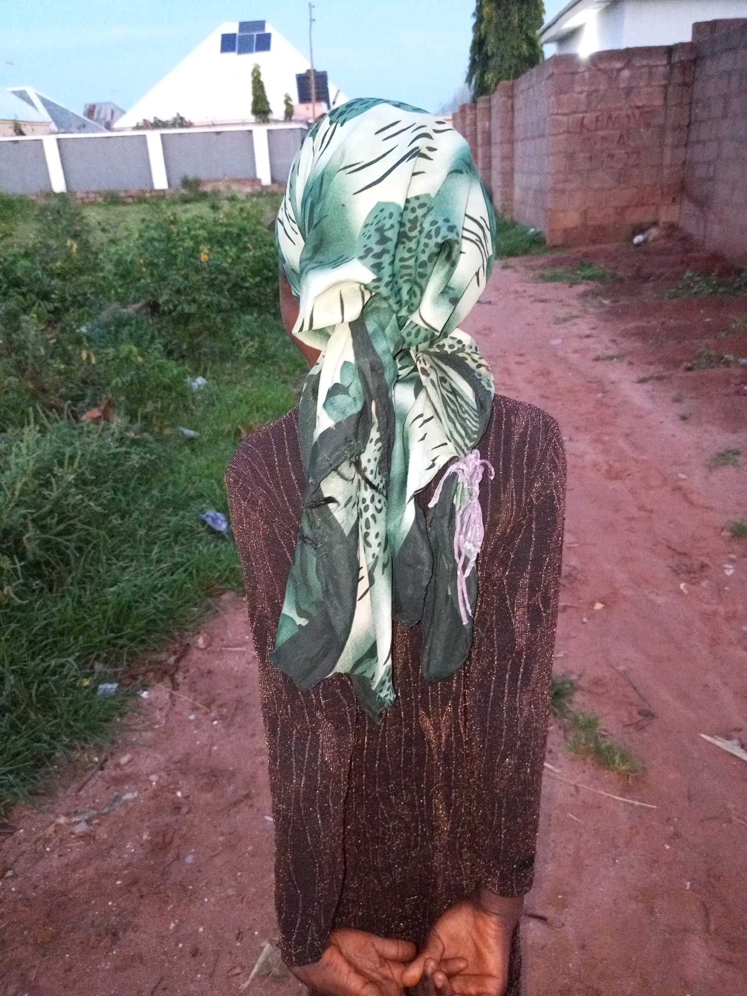 A full shot from behind shows a person wearing a brown, sparkly dress and a green and white patterned headscarf. The person's hands are clasped behind their back. The person is standing on a dirt path with green grass on the left side. In the background, there are buildings and a brick wall. The sky is light blue. The scene is located in the city of Tempure, Nigeria.