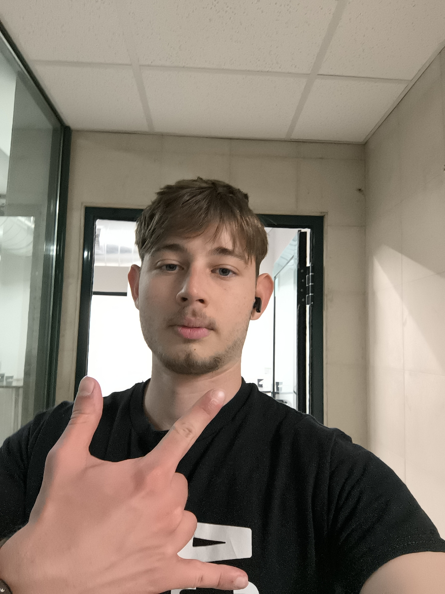 A young man takes a selfie indoors. He has light brown hair and blue eyes. He is wearing a black T-shirt and has an earbud in his right ear. He is making a hand gesture. The location appears to be a bathroom or utility area with a white tiled ceiling and walls. A window and a dark-framed door are visible in the background. There are no clear indications of this photo being in Sofia, Bulgaria.