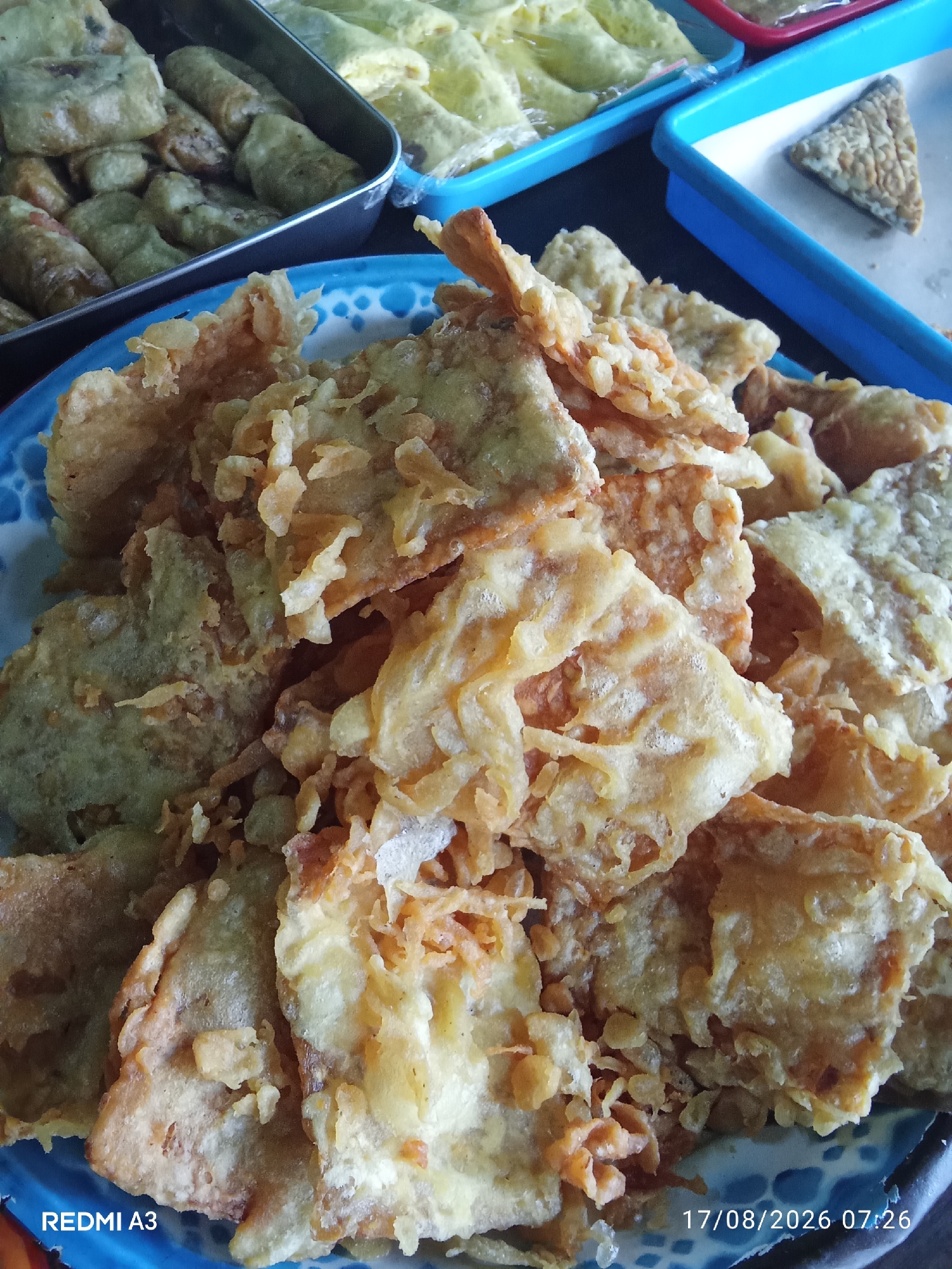 The image shows a close-up view of a plate of Indonesian fried food, likely tempeh chips or similar fried snacks, in Sanggrahan, Indonesia. The food is golden brown and appears crispy, piled high on a blue and white patterned plate. In the background, other trays of food are visible, including what look like spring rolls and other fried items. The lighting suggests an indoor setting, possibly a food stall or a home kitchen. The date stamp at the bottom right corner indicates "17/08/2026 07:26".