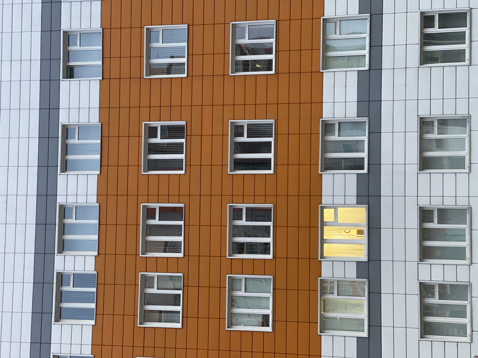 The image presents a close-up view of a modern building facade. The building has a distinctive design, featuring a combination of orange and white/gray vertical panels. The windows are uniformly square and white-framed, arranged in a grid-like pattern across the facade. Most windows have horizontal blinds or coverings partially obscuring the interior. Two windows on the right side show a yellow glow, indicating interior lighting. The facade appears to be tiled or paneled in a modular fashion. The scene suggests a contemporary urban architectural style. The location is not evident from the image; therefore, the city of Krasnoyarsk, Russia cannot be confirmed from this image alone.