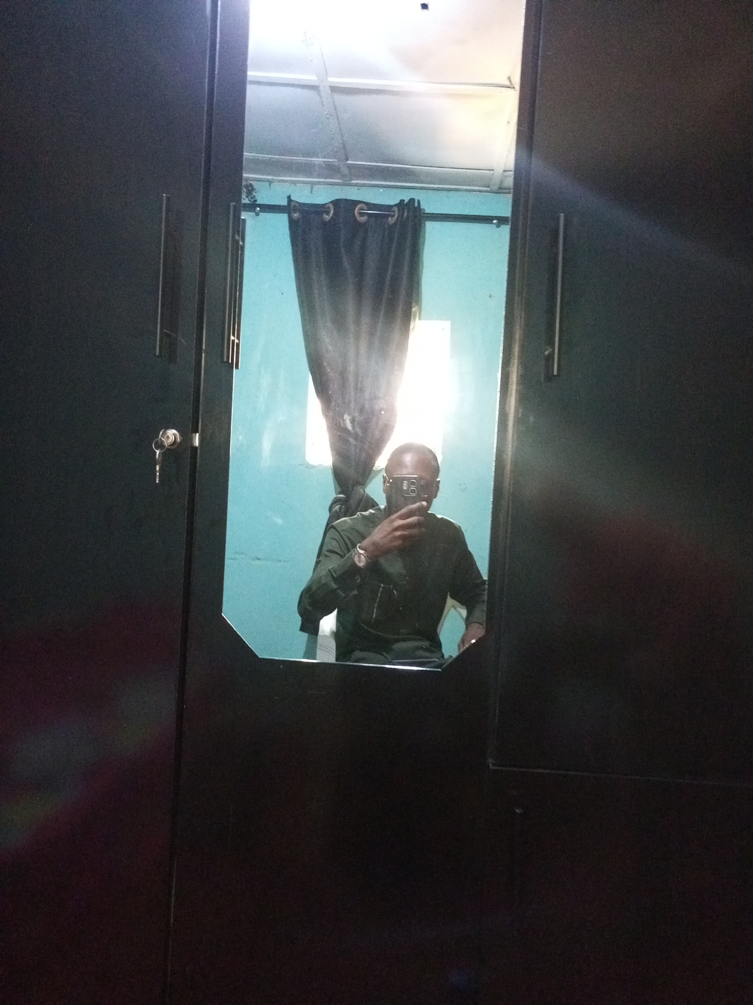 The image shows a self-portrait reflected in a dark-colored wardrobe mirror. In the reflection, a person wearing a dark green shirt and wristwatch is holding a mobile phone, presumably taking the picture. Behind the person is a blue wall partially covered by a dark curtain. A window lets in bright light, causing a glare in the reflection. A set of keys hangs from the wardrobe door. The location is Bukuru, Nigeria.