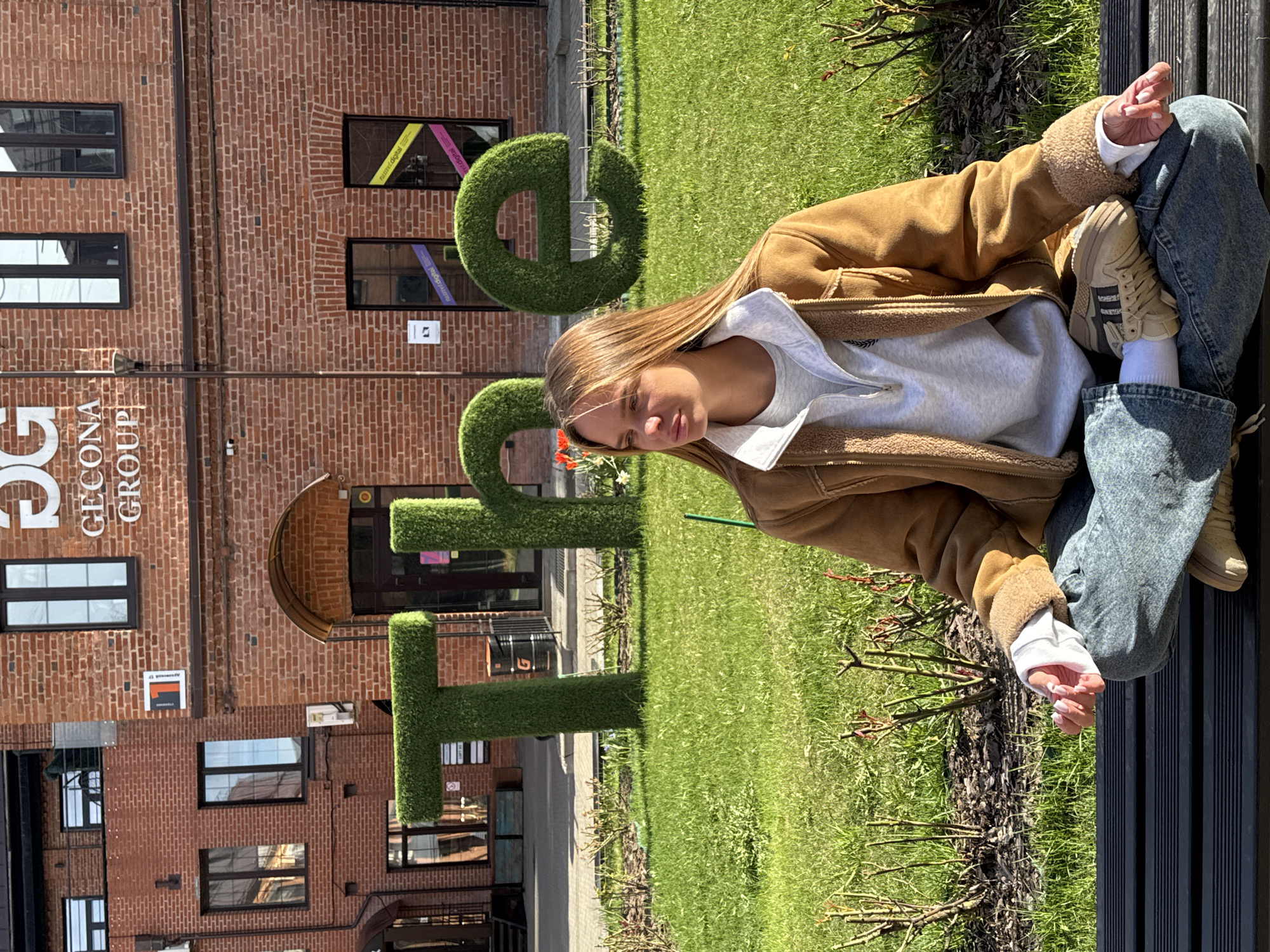 A young woman sits in a meditative posture on a black bench. She is wearing blue jeans, beige sneakers with black stripes, white socks, a gray sweatshirt, and a brown jacket with a sherpa lining. She is positioned with her legs crossed.
Behind her, an open green lawn leads to a building made of red bricks. The building has dark-framed windows, some of which have colorful decorations visible inside. In front of the building, topiary letters spell out "DGT" in green. A sign on the building reads "GEOCONA GROUP."
The location is Moscow, Russia.