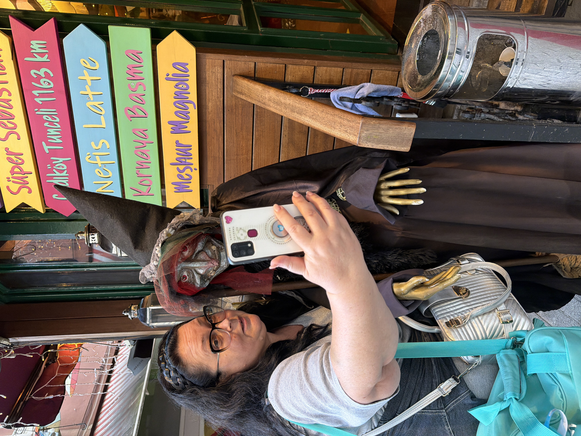 A woman with black hair and glasses is taking a photo with her white smartphone of a Halloween witch display in what appears to be a storefront window. The witch figure has a black hat, a silver mask, and gold-colored hands protruding from a brown robe. Wooden signs point toward different locations, including "Süper Sebastia," "Çalköy Tunceli 1163 km," "Nefis Latte," "Kornaya Basma," and "Meşhur Magnolia." A silver ashtray is located above the witch figure. The scene is outdoors, and the photo is taken at eye level, likely in the city of Kadıköy, Turkey.