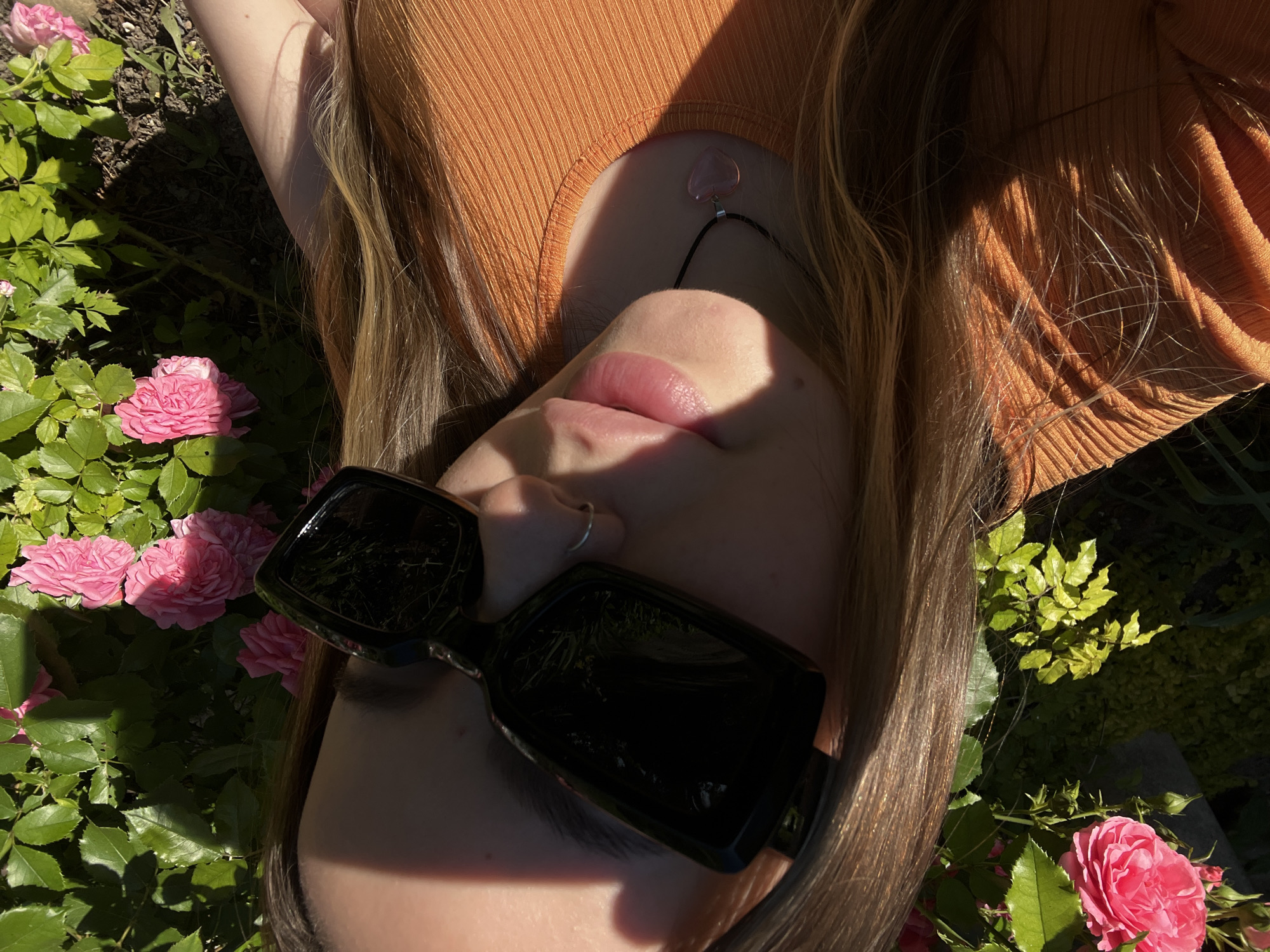 A young woman with long, light brown hair is captured in a close-up, outdoor selfie. She is wearing large, black, rectangular sunglasses and an orange ribbed top. A thin, silver-toned necklace with a light pink heart-shaped pendant is visible against her top. She has a silver hoop nose ring. The background consists of lush green foliage and several pink roses, suggesting a garden setting. Sunlight casts dappled shadows across the scene, highlighting the textures of the leaves and the woman's hair and clothing. The image appears to have been taken during the daytime. The location is identified as the city of Novopokrovskaya, Russia.
