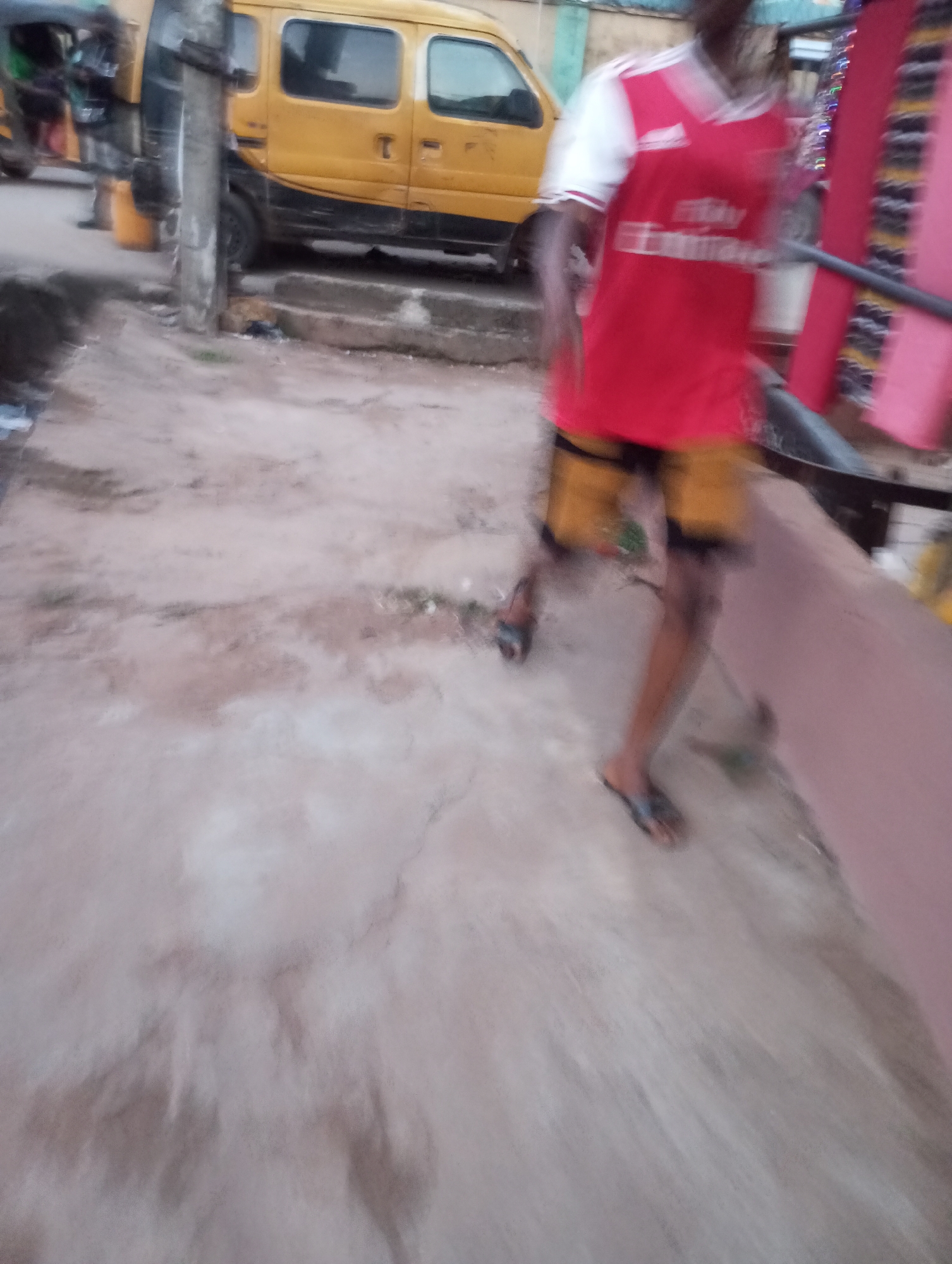 The image captures an outdoor street scene in what appears to be an urban or semi-urban area, consistent with Onitsha, Nigeria. A person is depicted in motion, walking from left to right across the foreground, causing significant blur. They are wearing a red and white short-sleeved football jersey, identifiable as an Arsenal F.C. kit by the red body, white sleeves, and visible Adidas logo and "Emirates Fly Better" sponsor text. They also wear yellow or light brown shorts and black flip-flops.
In the midground, to the left of the moving person, a yellow panel van or minibus, possibly a commercial passenger vehicle, is parked or stopped. A utility pole stands directly in front of the van, partially obscuring it. Further to the left and slightly behind, a green and black three-wheeled auto-rickshaw (often called a 'keke Napep' in Nigeria) is partially visible. A yellow bucket is placed on the ground near the tricycle.
The ground surface consists of concrete or paved earth, showing signs of wear and discoloration. A raised curb or step separates the paved area from what seems to be a road or unpaved section. On the far right, a section of a pink building or structure is visible, featuring some decorative elements, possibly string lights. The lighting is soft and ambient, suggesting it is daytime but possibly overcast, early morning, or late afternoon. No specific emotions are discernible due to the blur and distance.