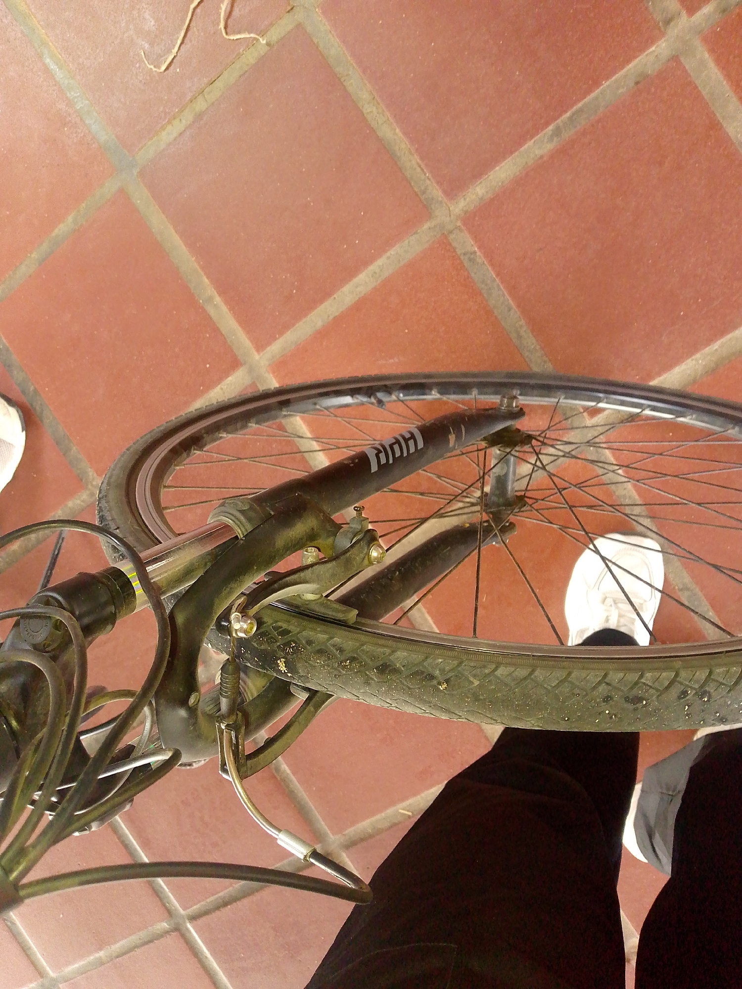 A partial overhead view captures a bicycle’s front wheel resting on red, square floor tiles. The tire appears used, with visible wear. The bicycle frame is dark, possibly black, and partly obscured. A leg clad in black pants and a white sneaker is positioned near the wheel. A second white sneaker is located behind the wheel. The location appears to be indoors. The scene is not conclusively linked to Pedro González, Venezuela.