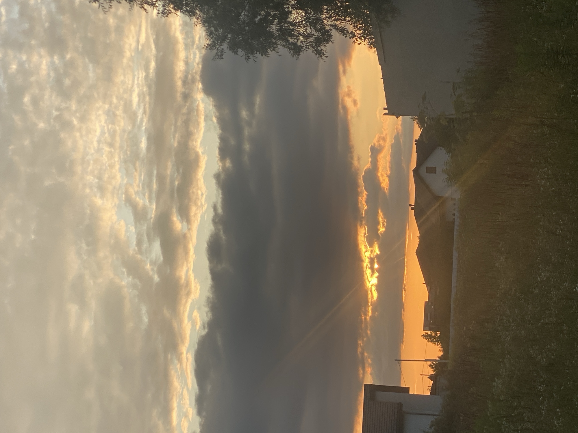 This is a vertical, eye-level, medium shot of a sunset over a rural area. The sky is filled with a mix of dark and light clouds, with beams of light breaking through the clouds, illuminating the sky with golden and orange hues. In the foreground, there is a grassy field with tall weeds and some small white flowers. In the mid-ground, there are several houses with tiled roofs, a utility pole, and some trees. A large, dark building is visible on the right side of the frame. The overall impression is a serene evening in a suburban or rural setting. The specific location is identified as Novi Petrivtsi, Ukraine. There are no people or animals visible in the image.
