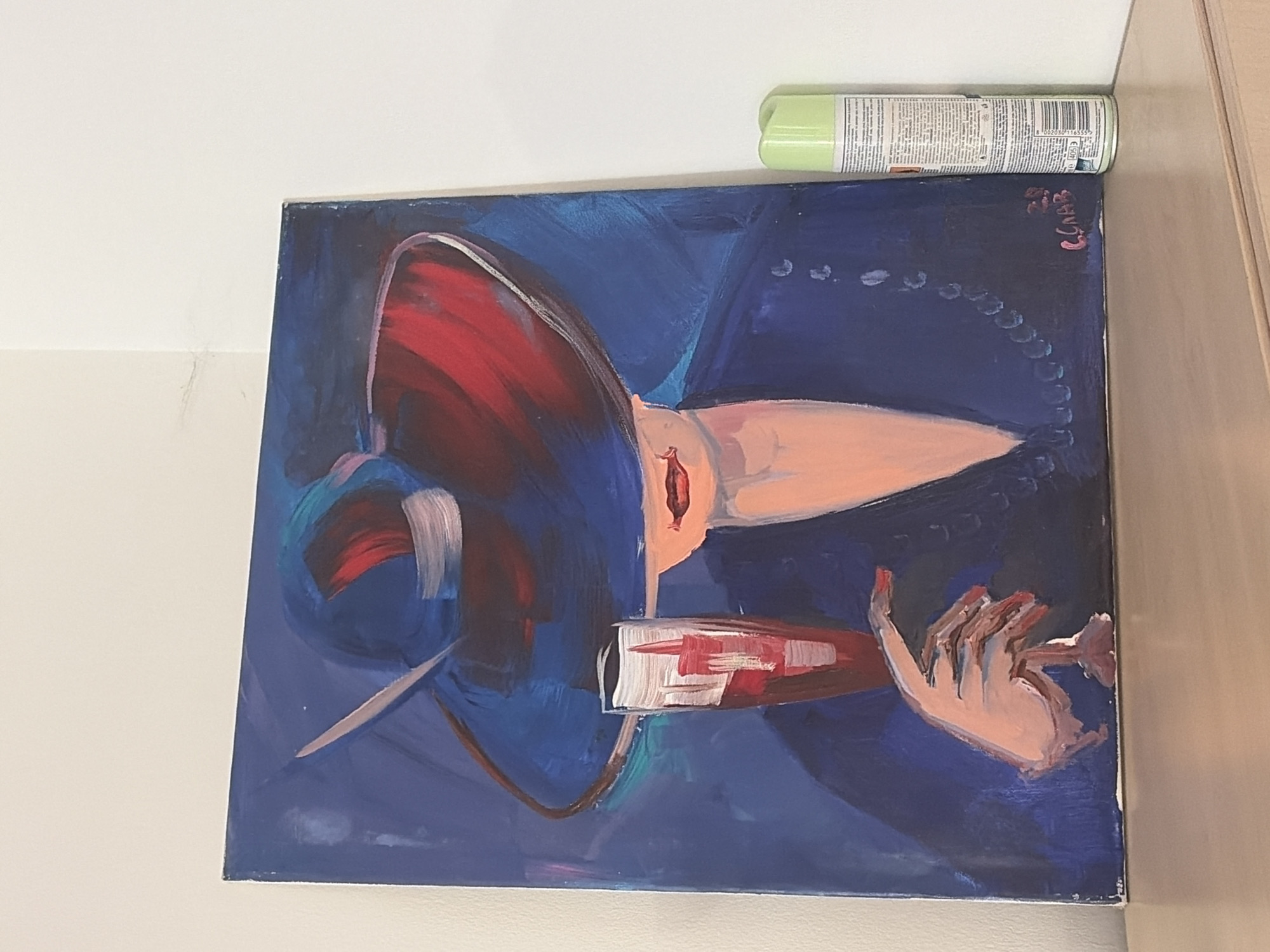 A painting dominates the frame, depicting a stylized portrait of a woman in a blue dress and a red hat. She holds a white and red object. A string of pearls loops around her. Above the painting sits a can of green spray paint. The environment is an interior space, with a light-colored wall visible to the left and a beige or light brown door or cabinet on the right. The city of Sofia, Bulgaria, is not discernible from the image.