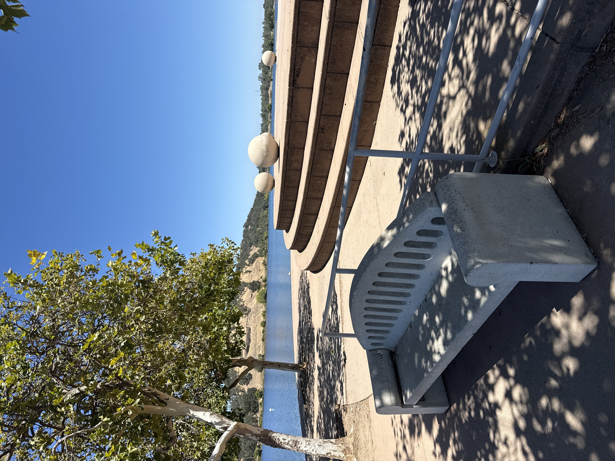 A serene outdoor scene unfolds at Hagan Community Park in Rancho Cordova, California, under a clear blue sky. A concrete bench, weathered by time and dappled with sunlight filtering through overhead trees, sits in the foreground. Beyond the bench, a tiered, circular amphitheater-like structure made of stone steps rises, topped with three large, smooth, spherical sculptures. The lake serves as a backdrop, with hills dotted with trees lining the opposite shore. A lone tree with a mottled white trunk stands to the left, its branches reaching towards the sky. The overall impression is one of tranquility and natural beauty.