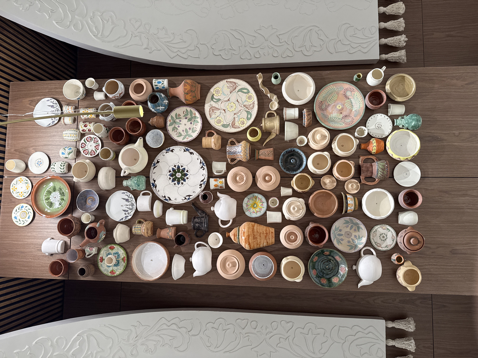 The image shows a collection of ceramic vessels arranged on a dark brown, horizontally-grained surface, possibly a table. The collection includes plates, bowls, cups, mugs, pitchers, and pots of varying sizes, shapes, and decorative patterns. Many of the items appear to be handmade or artisan pieces. Above and below the collection are parts of white, decorative panels with floral patterns and tassels. The location is presumed to be in Kazan, Russia, given the visual details of the crafts. No people are visible in the frame. The objects are still and appear to be staged or arranged for display purposes. The lighting is relatively bright, casting soft shadows.