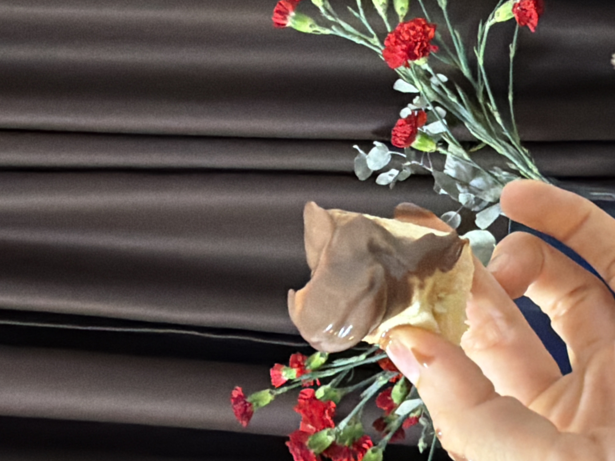 An indoor, close-up shot captured in Saint Petersburg, Russia, features a human hand holding a piece of a chocolate-dipped dessert against a dark, draped fabric background. The dessert appears to be a light-colored pastry, possibly a cream puff or choux pastry, partially covered with rich, melted milk chocolate that glistens slightly. The hand, with neatly manicured fingernails, delicately grasps the dessert.
In the background, positioned to the right and subtly extending towards the upper left, is a bouquet of vibrant red flowers, resembling miniature carnations, interspersed with slender green stems and lighter green or greyish foliage, possibly eucalyptus. The dark background fabric, likely satin or velvet, is arranged in soft, horizontal folds, suggesting a curtain or tablecloth. The lighting is soft and diffused, creating a gentle focus on the hand and dessert, and highlighting the textures of the various elements. There is no visible text in the image.