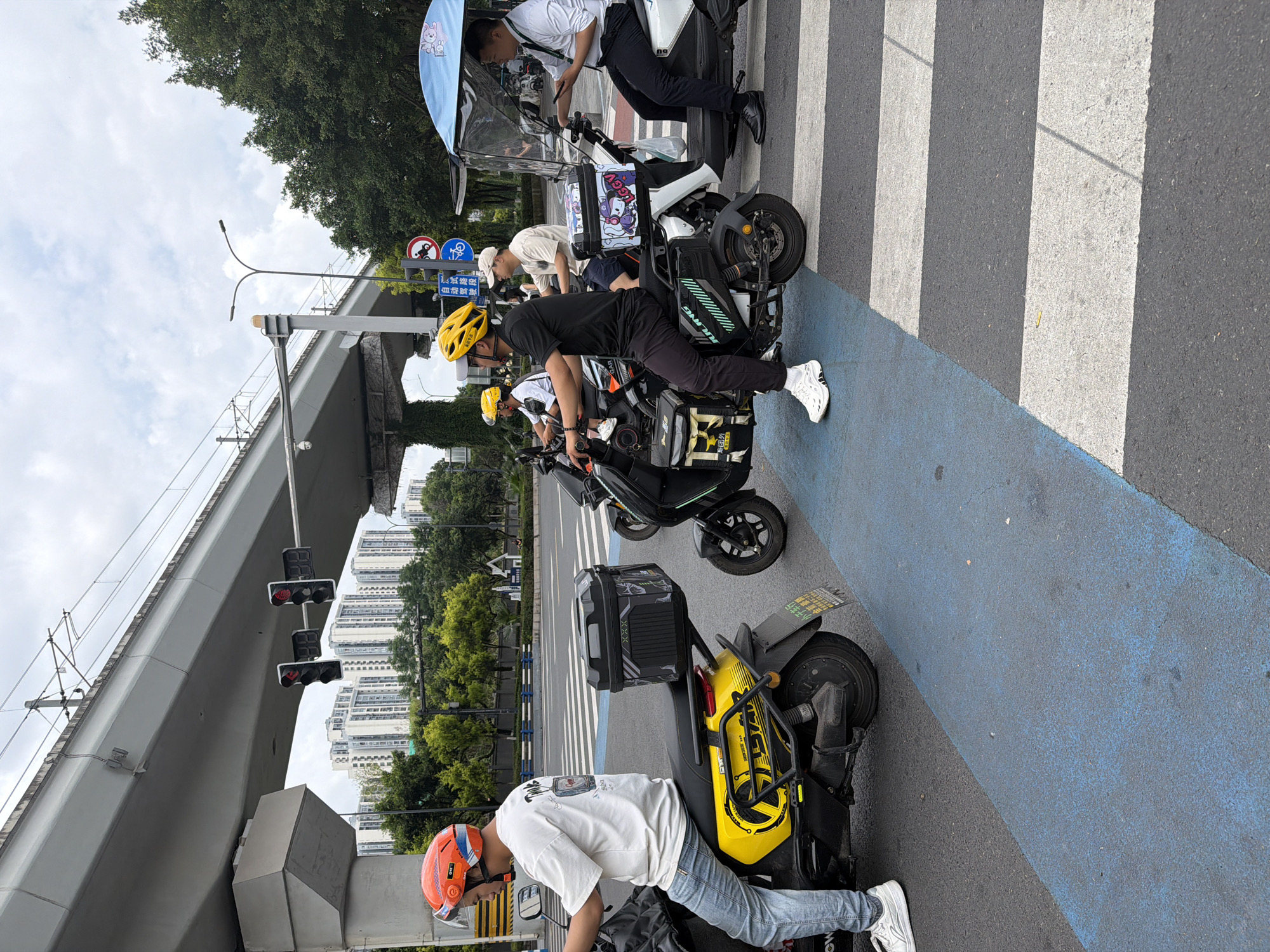 The image depicts a street scene in Chongqing High-tech Zone, China. Four people are straddling electric scooters or motorbikes at a crosswalk. Two of the scooters are white, one is black and one is yellow. One of the riders wears a white shirt, jeans, a bright orange helmet. Another wears a black shirt, black pants, a yellow helmet and white shoes. The scooter in front of him is black, and has a delivery bag attached. One rider sits under a light blue canopy. An elevated section of road is over head, and a pedestrian crossing sign is visible on a lamp post. The street is marked with blue lines along the road edge and in front of the crosswalk. Traffic lights are visible above the elevated section of road, and a building is in the background.