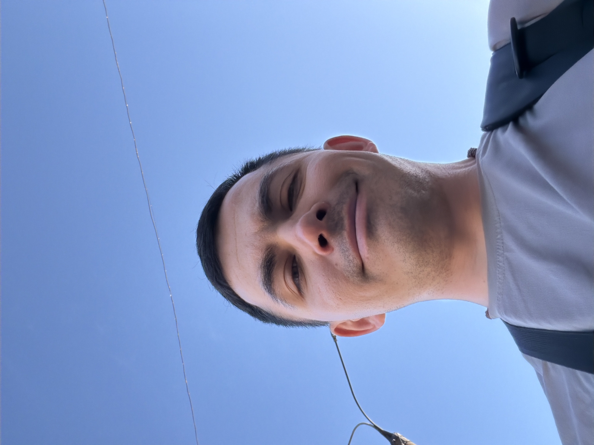 A man with dark hair and light skin is pictured from a low angle, looking down at the camera. He has a slight smile. He is wearing a light grey shirt and what appears to be a backpack. The background is a clear blue sky with a power line visible in the upper left. The location is not discernible from the provided image.