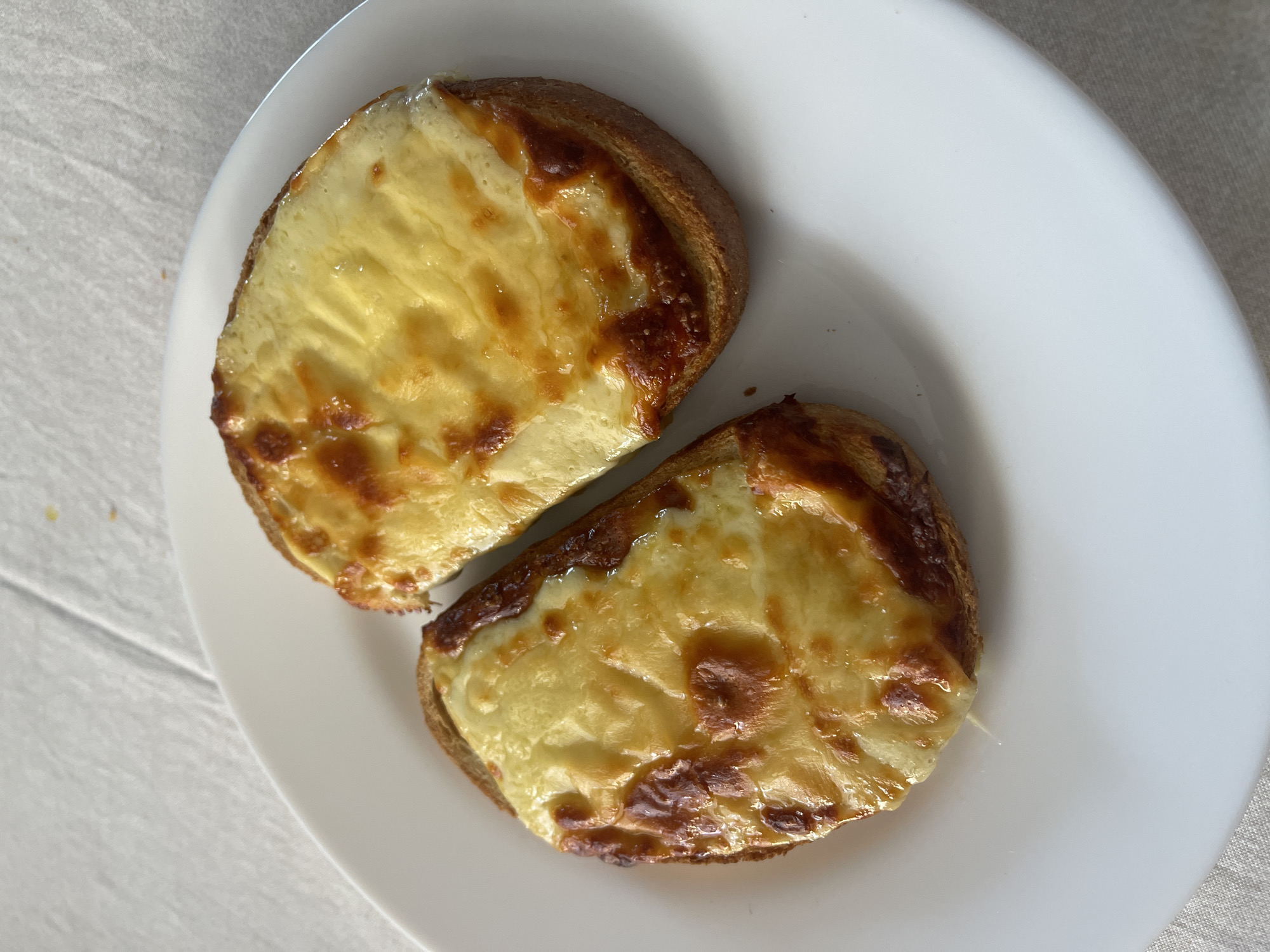 The image shows a white plate with two slices of bread topped with melted cheese. The cheese is golden brown in places, indicating it has been heated. The bread appears toasted. The plate is sitting on a surface that includes wood and a textured cloth. The location is Unknown, Unknown. There are no people in the image.