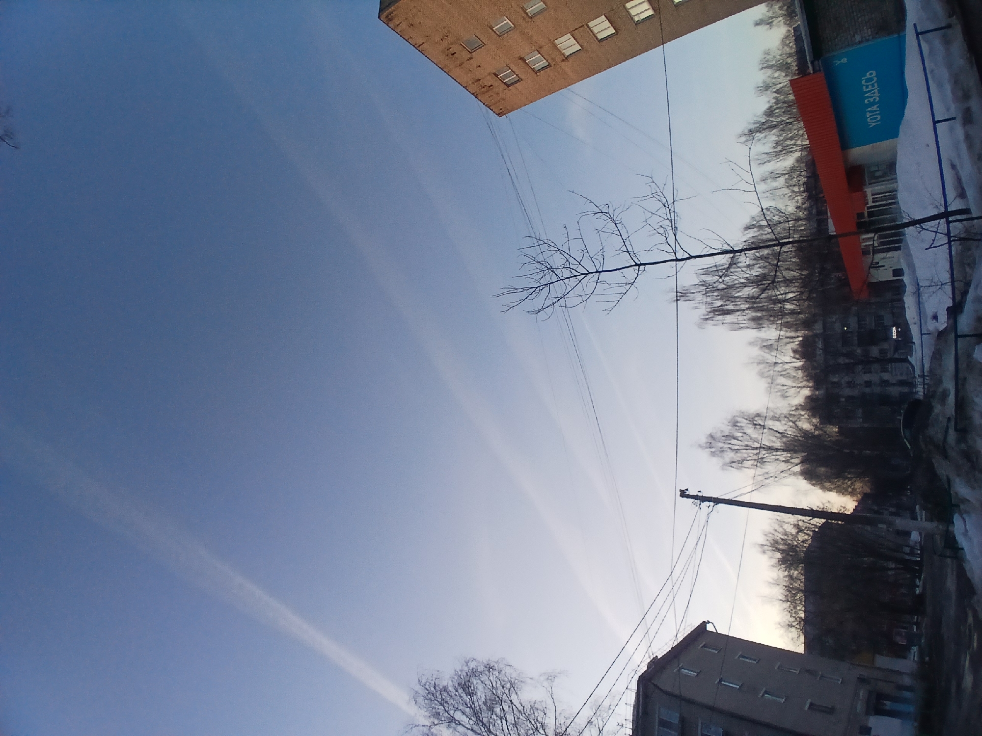 The image captures an urban scene in what appears to be Chaikovsky, Russia, judging by the “YOTA ZDEs’" sign partially visible on a building. The photograph is taken from a low angle, tilting upwards to emphasize the sky. The sky occupies a significant portion of the frame and exhibits streaks, possibly contrails. Several buildings are visible, including a brick building in the upper left, a building with the YOTA sign, and a smaller residential building in the lower right. Power lines crisscross the frame, adding to the urban aesthetic. Bare trees are interspersed among the buildings. The right side of the image shows snow-covered areas.