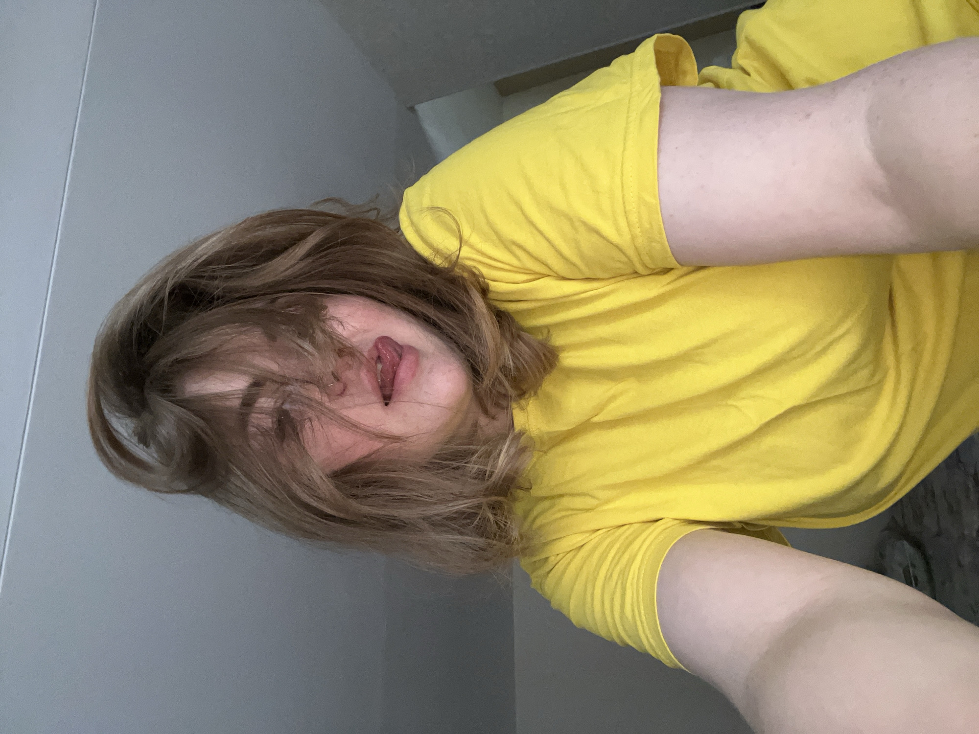 A woman with brown hair is wearing a yellow t-shirt. She is sticking her tongue out. The location is unknown.