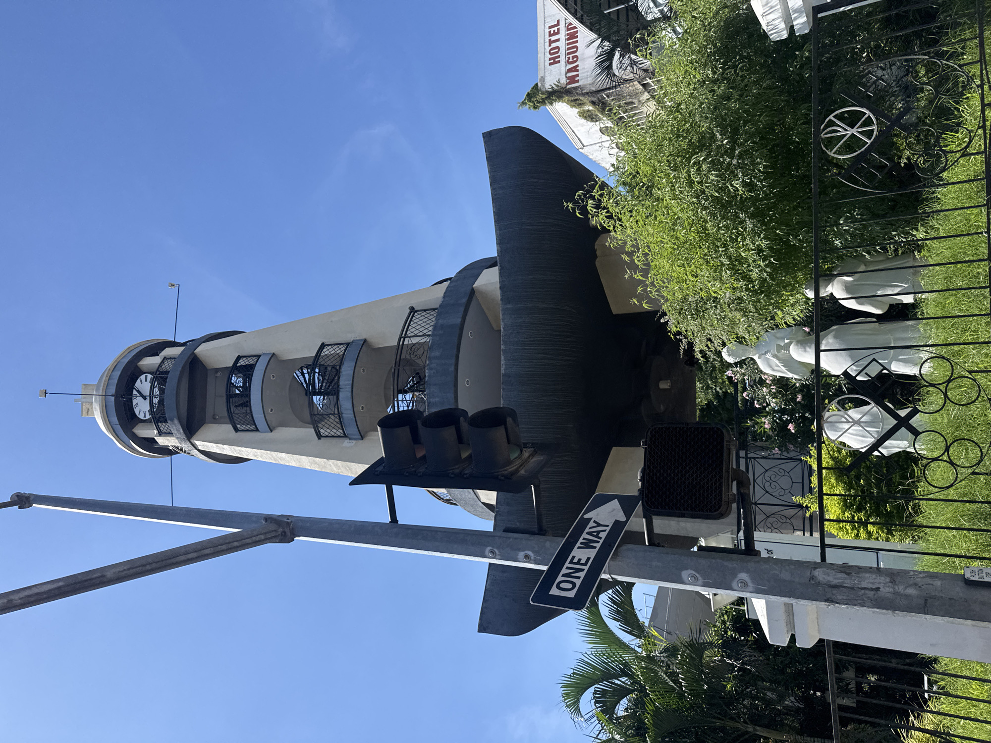A low-angle, full-shot photograph captures a segment of the iconic Clock Tower of the Insular Life Building in Davao City, Philippines, under a clear blue sky. The tower, an architectural landmark, stands tall with its distinctive beige facade, multiple balconies with ornate railings, and a clock face visible at its apex. Below the tower, a large, dark, boat-like structure juts out, adding a unique design element.
In the foreground, a traffic light pole with a "ONE WAY" sign pointing left is prominently positioned. The traffic light itself is dark and appears to be a standard three-light signal. To the right of the pole, a pedestrian crossing signal box is visible. Behind the traffic light pole, lush green foliage and palm trees add a tropical feel to the scene.
Further to the right, a portion of "HOTEL MAGUIOIND" is visible, indicating the proximity of this establishment. In front of the hotel and peeking through the greenery are white statues, possibly religious figures, standing behind a black wrought-iron fence. One statue, taller than the others, is positioned in the center, and two smaller ones are to its right. The fence has a circular emblem with a cross-like symbol inside. The ground is covered in green grass. The overall impression is a sunny day in a city that blends modern architecture with natural elements and cultural markers.