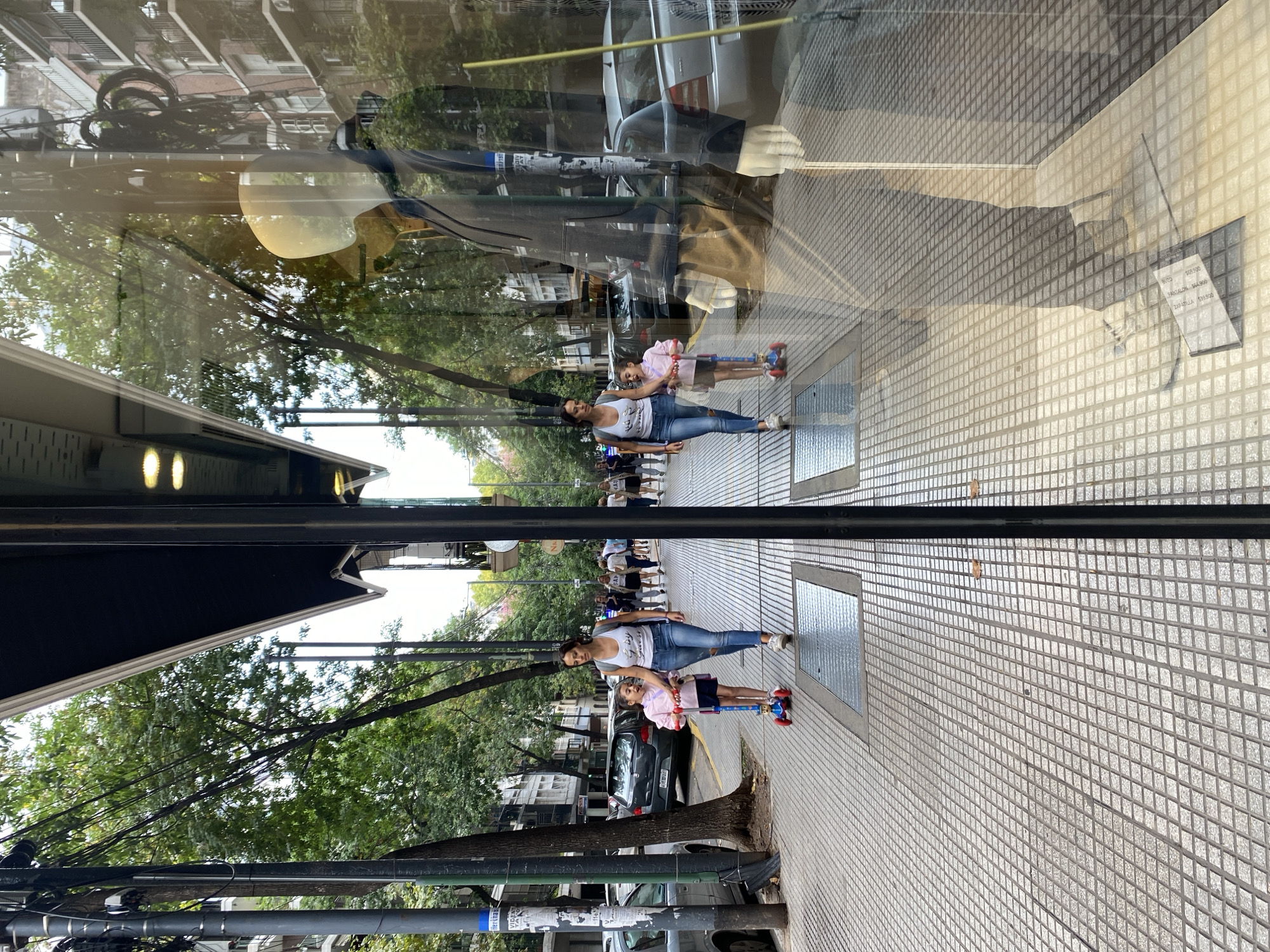 The photograph captures a daytime urban street scene in Buenos Aires, Argentina, primarily through the reflective surface of a storefront window.
In the foreground, a prominent vertical black structural element, likely part of the window frame or building facade, divides the central-right portion of the image. The ground consists of a gray sidewalk paved with a repeating diamond-grid pattern, featuring two recessed rectangular metal grates. The reflection of a mannequin is visible within the window's right side, showing its translucent head and white hands, clad in a dark jacket. A small, partially obscured white sign with text, including "PANTALON" and "REMERA" with associated prices, lies on the reflected floor.
The reflected street scene depicts a woman and a young girl walking from left to right on the sidewalk. The woman, on the right, wears a white tank top with a dark graphic, light blue ripped jeans, and carries a dark backpack. She holds the left hand of the girl beside her. The girl wears a light pink long-sleeved shirt, dark shorts, and rides a red and blue kick scooter. The sidewalk is lined with numerous mature trees featuring dense green foliage. Utility poles with complex networks of black overhead wires are visible on the left. Several parked vehicles, including a dark hatchback and a silver sedan, line the street. Multi-story buildings, characteristic of urban residential and commercial structures, extend into the background.
Additional pedestrians are visible further down the street, primarily on the right side. The sky appears bright and overcast.