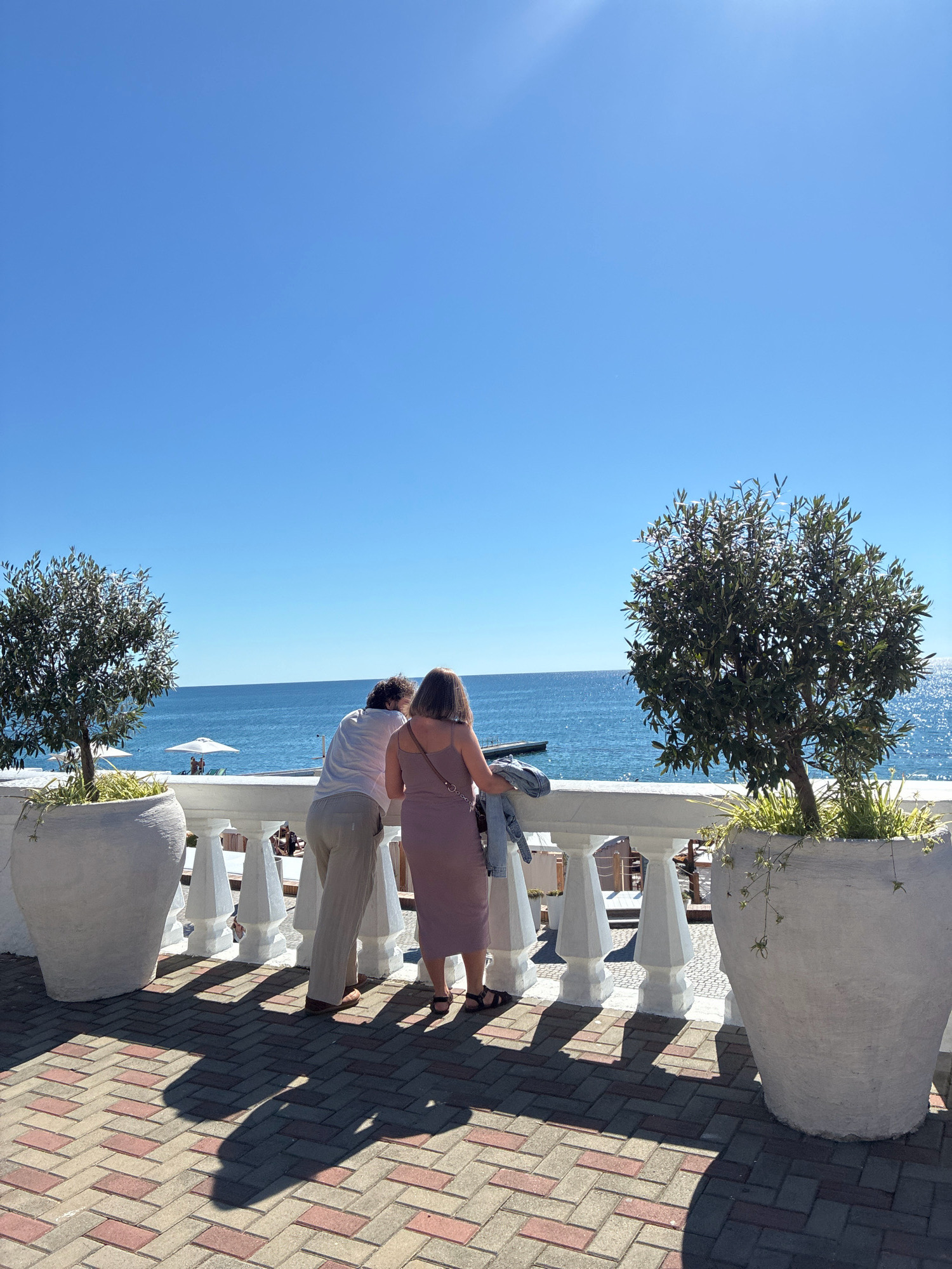 Two individuals, a man and a woman, stand at a white railing overlooking the Black Sea. The man, wearing a white shirt and beige pants, leans on the railing. The woman, in a pink dress, stands beside him, holding a denim jacket over her arm. They are on a tiled patio with large white planters containing small trees on either side. The sea is calm and blue under a clear sky. In the distance, beach umbrellas and people are visible on the shore. The sun casts distinct shadows on the patterned tiles. The scene depicts a serene moment by the sea, likely at a cafe or restaurant terrace along the Sochi coastline.