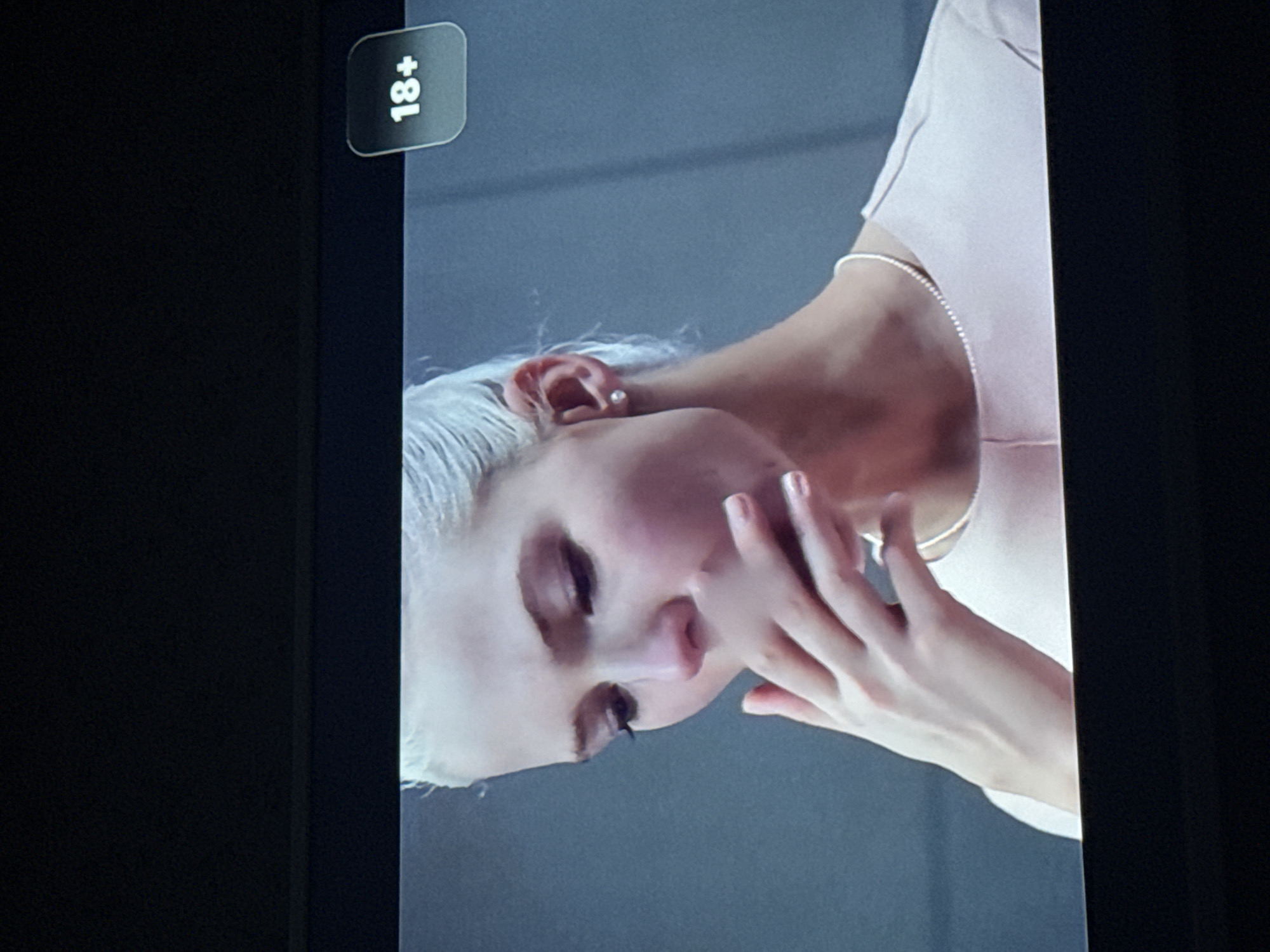 A close-up of a person with white hair, fair skin, and a light pink shirt. The person's face is tilted downwards and to the left, with the eyes closed and a hand touching the face. An "18+" rating label is visible in the upper left corner. The background is a plain gray color. The location is unknown; the information provided is not sufficient to determine if it is in Mytishchi, Russia.