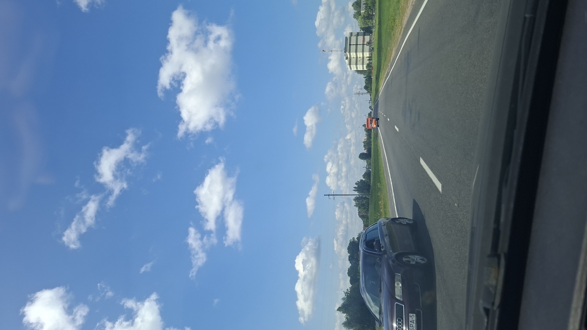 An outdoor, medium shot captures a view from a moving vehicle looking down a paved, two-lane road under a bright blue sky with scattered white clouds. On the left side of the road, a dark red station wagon is in motion. On the right side of the road, an orange truck is also moving forward, with a multi-story building in the distance. The environment appears to be rural, with grassy fields and trees lining the sides of the road. A power line pole is visible near the left side of the road. The perspective suggests the image was taken during the daytime.