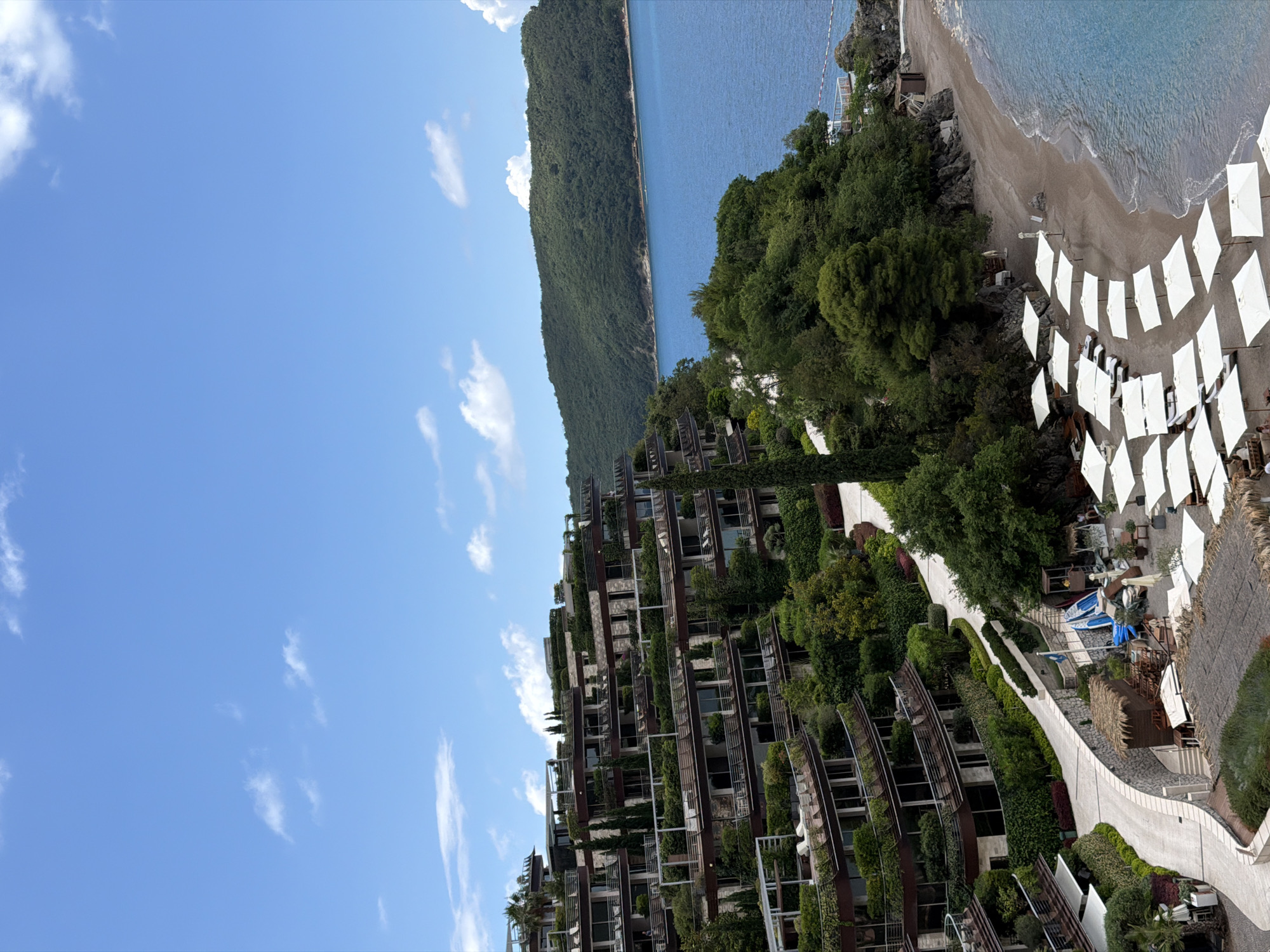This vertically oriented image captures a picturesque coastal resort scene in Boreti, Montenegro, under a clear blue sky with scattered white clouds. The setting features a modern, terraced building seamlessly integrated into a lush green hillside, overlooking the Adriatic Sea and a sandy beach.
The central subject is a multi-story building, likely a luxury hotel or residential complex, characterized by its distinctive architecture with numerous balconies and extensive vertical gardens and planted greenery that cascade down its façade. This design allows it to blend harmoniously with the surrounding natural landscape.
To the right of the building, a pristine sandy beach stretches along the coastline, dotted with numerous neatly arranged white sun umbrellas and matching lounge chairs, suggesting a well-appointed resort beach ready for guests. Gentle waves lap at the shore, and the clear blue-green water of the sea extends into the distance. A few small boats, possibly kayaks, are visible near some beachside structures.
In the background, a long, verdant mountain range covered in dense forest extends along the horizon, creating a dramatic backdrop to the tranquil sea. Winding paved pathways traverse the resort grounds, bordered by manicured hedges and abundant trees and shrubs, leading guests through the landscaped areas. Some small, thatched-roof structures can also be seen near the pathways and beach, likely offering amenities or dining options.
The scene depicts a calm, sunny day, probably late morning or early afternoon, with bright illumination. The overall impression is one of luxury, relaxation, and natural beauty. There is no visible text in the image, and no specific activities or people are distinctly visible, emphasizing the serene ambiance of the resort.