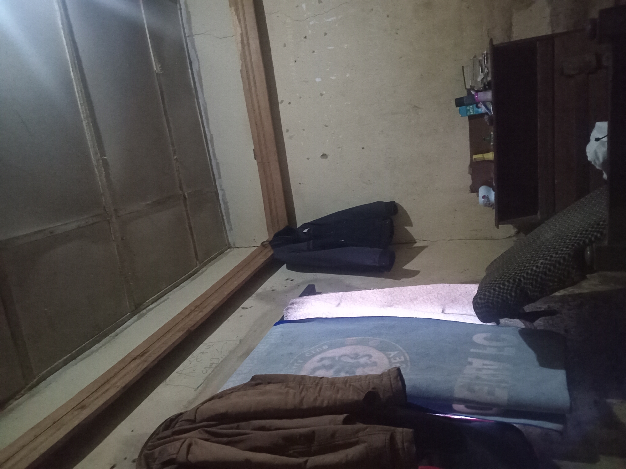 An indoor scene with rough, unpainted walls and a ceiling constructed of gray panels with visible wooden beams. Two jackets are hanging on the wall, one brown and one dark blue. A large blue cloth with white lettering is also hanging, partially obscured by the jackets. A wooden shelf unit is against the wall, holding various items including bottles and papers. In the foreground, part of a patterned cushion or chair is visible. The lighting suggests artificial light sources.