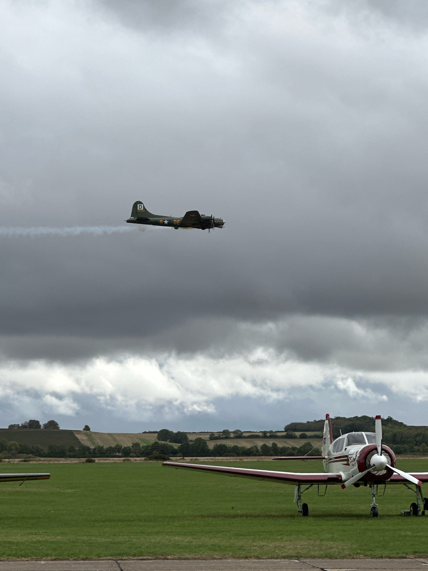 A B-17 Flying Fortress aircraft is in flight under a cloudy sky, leaving a smoke trail. In the foreground, a vintage propeller-driven airplane is parked on a grassy field. The background shows rolling hills with agricultural fields and trees. The scene is likely at an airshow or airfield in South Cambridgeshire, United Kingdom.