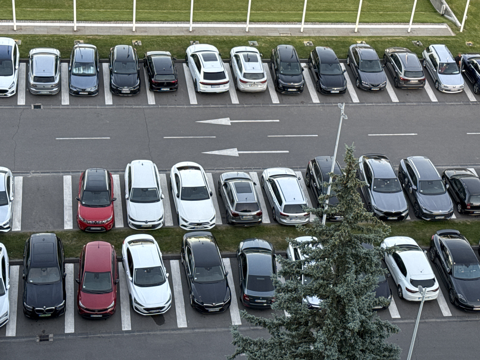 The image depicts an aerial view of a parking lot in Unknown, Unknown. Two rows of parked cars are visible. A road with white directional arrows separates the two rows of vehicles. A tree is positioned in the lower right corner of the frame, obscuring some of the vehicles. Grass and white posts line the top edge of the frame. The image suggests an urban or suburban setting with parking facilities.