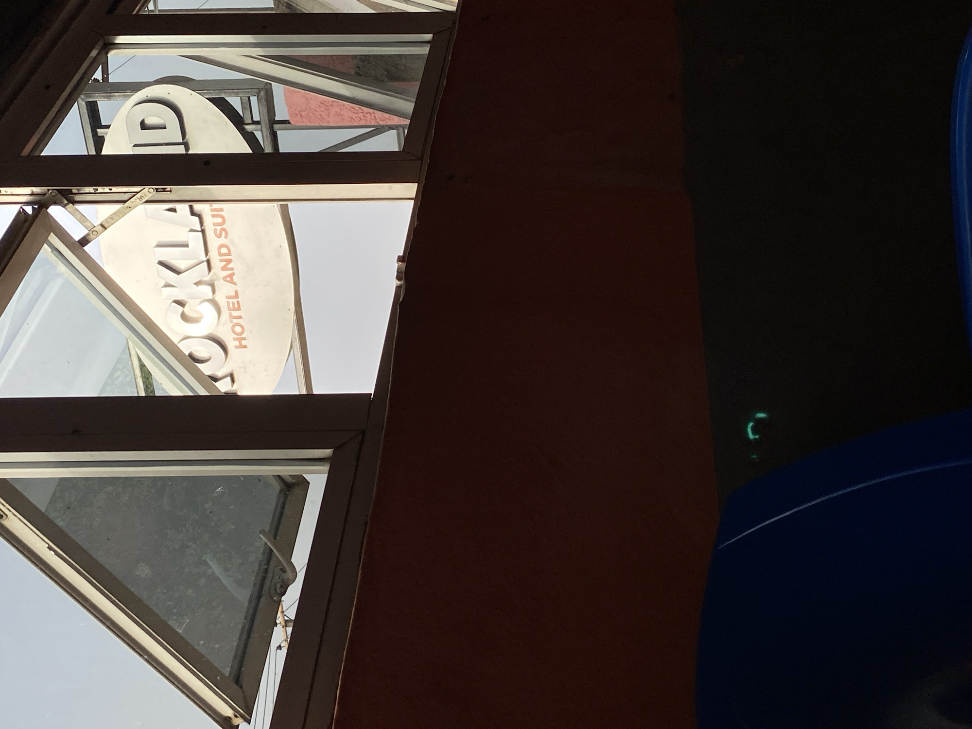 The image, taken in Jos, Nigeria, presents a view from what appears to be an interior space, looking outwards through windows towards an illuminated exterior. The left side prominently features multiple dark-framed windows or angled glass panes.
Through these windows, a large, light-colored sign for a business is clearly visible. The upper portion of the sign displays the bold, dark letters "LD." Below this, the larger text "ROCKLA" is seen, strongly suggesting "ROCKLAND." Further down, smaller, reddish-brown lettering spells out "HOTEL AND SUI," indicating "HOTEL AND SUITES." Metal support structures are visible around the sign. The bright illumination on the sign suggests it is daytime outside.
The right side of the image is considerably darker, dominated by a large, reddish-brown wall or architectural element that extends into a very dark, almost black area. In the bottom right corner of this dark section, a curved, bright blue object, possibly part of a chair or a plastic container, is discernible. Just above this blue object, a small, faint green light, shaped like an incomplete 'C' or bracket, glows in the darkness, hinting at an electronic device or indicator.
No people are present, and the scene is static, capturing a moment of stark contrast between the brightly lit exterior sign and the shadowed interior space from which the photo was taken.