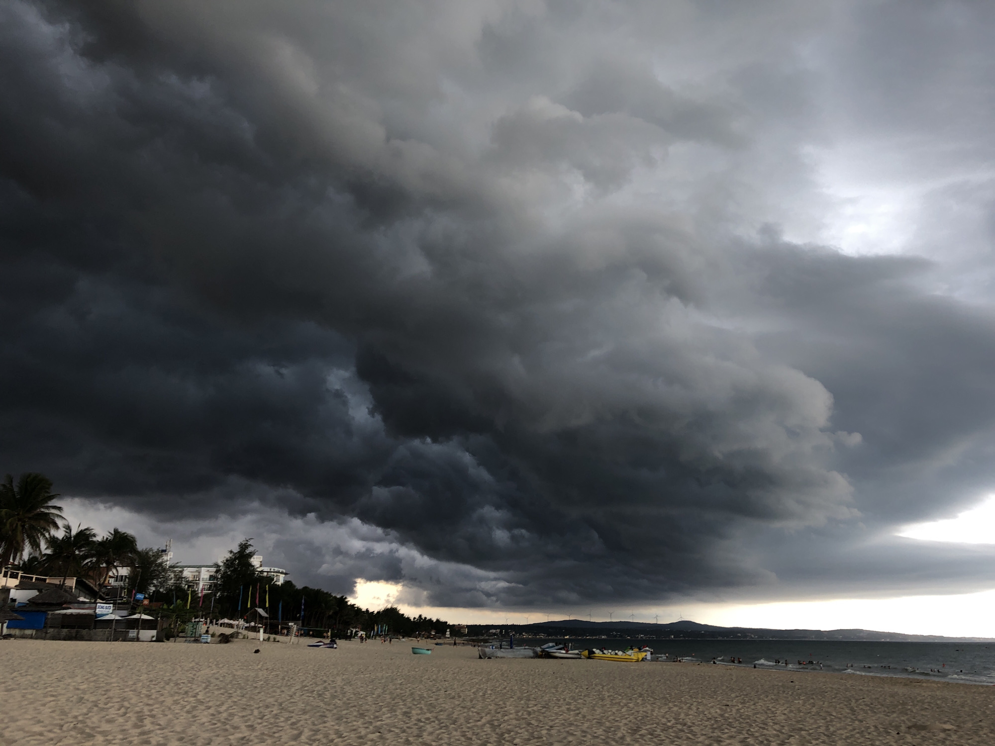 A wide shot captures a beach in Mũi Né, Vietnam, under a dramatic sky filled with dark, heavy storm clouds that dominate the upper two-thirds of the frame. The clouds are a mix of deep grays and blacks, suggesting an impending downpour. The lower portion of the image shows a sandy beach leading to the ocean. Along the shoreline, a few palm trees are visible on the left, interspersed with small buildings and flags. Several small boats are pulled up onto the sand, and a few people are present on the beach and in the water. The ocean itself is a muted gray, reflecting the overcast conditions. In the distance, a hazy coastline with some structures and what appear to be wind turbines can be seen. The overall atmosphere is ominous due to the threatening weather.
