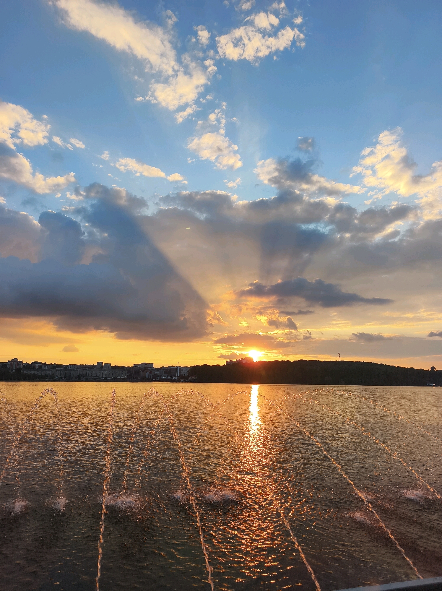 The image depicts a sunset over a body of water, likely Ternopil Lake, in Ternopil, Ukraine. The sky is a dramatic mix of blue and orange, with rays of sunlight piercing through dark clouds. The sun is low on the horizon, casting a golden reflection on the water's surface. In the foreground, several fountains spray water, creating an arc of droplets that catch the light. In the distance, the cityscape of Ternopil is visible, with buildings lining the shore, and a dark, tree-covered hill forms the far bank of the lake. The scene is tranquil and evocative of the end of a day. There are no people or animals visible in the image.