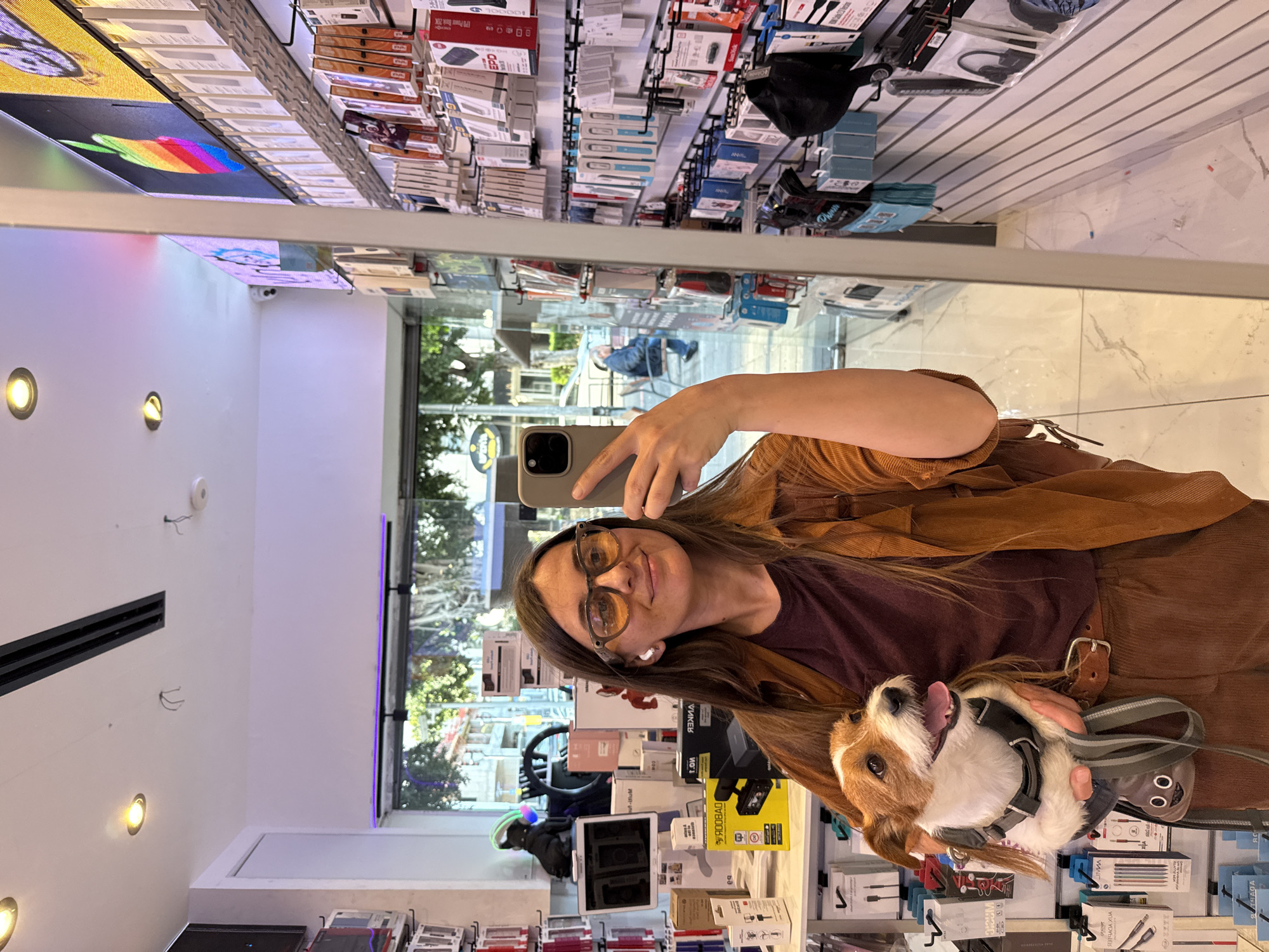 A woman is taking a selfie with her dog in a store, reflected in a mirror. The woman is wearing glasses, has brown hair, and is wearing a brown corduroy jacket. She is holding a smartphone in her right hand. The dog is small and brown and white, wearing a harness.
The store appears to sell electronics accessories, as shelves are lined with boxed items, possibly phone cases, headphones, and chargers. Through the window reflected in the mirror, a street scene is visible. There are trees and buildings. A person is sitting in a chair outside.
The location is Raanana, Israel, based on the streetscape visible in the reflection.