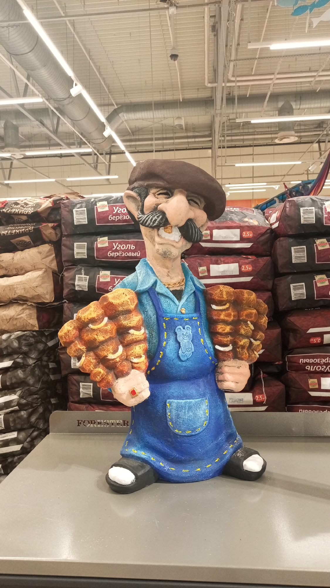 The image shows a whimsical, painted garden gnome-like figurine of a baker or peddler. He's wearing a brown beret, a blue shirt, and a blue apron adorned with a teddy bear design and yellow stitching. He has a mustache and is holding what appears to be clusters of baked goods, possibly pretzels or bread rolls, in each hand. He also sports a ring with a red stone. The gnome is placed on a gray surface with the word "FORESTER" on it. Behind the figurine are stacks of bagged charcoal, some labeled in Russian. The location appears to be a store, likely a gardening or home improvement center, with industrial-style ceiling fixtures and vents visible. The presence of Russian text suggests the location is in Russia. The Yudino location detail is not evident from this image.