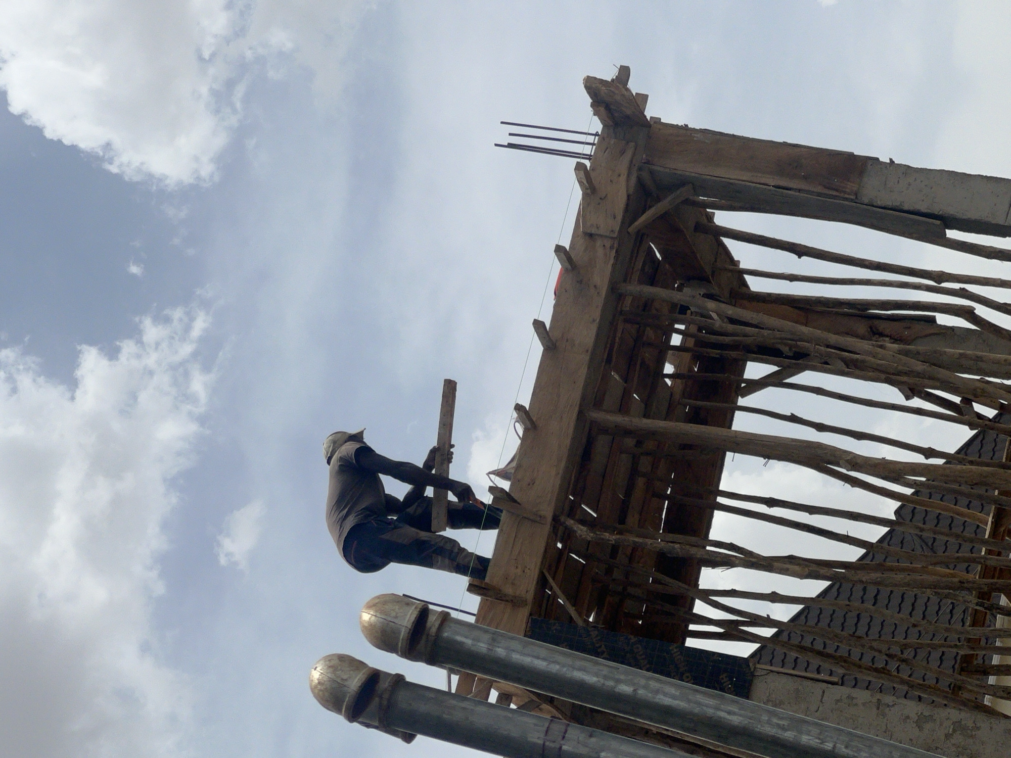 A low-angle shot depicts a construction worker on a wooden structure against a cloudy sky. The worker, wearing dark clothing and a cap, appears to be attaching a wooden plank to the structure, possibly with a hammer or similar tool. The wooden structure is complex, featuring a framework of beams and branches. In the foreground, three vertical silver pipes are visible. This construction site is likely located in Jos, Nigeria, as indicated by the local architectural practices with the use of wood and branches in building. The shot’s angle emphasizes the height and precarious nature of the construction work.