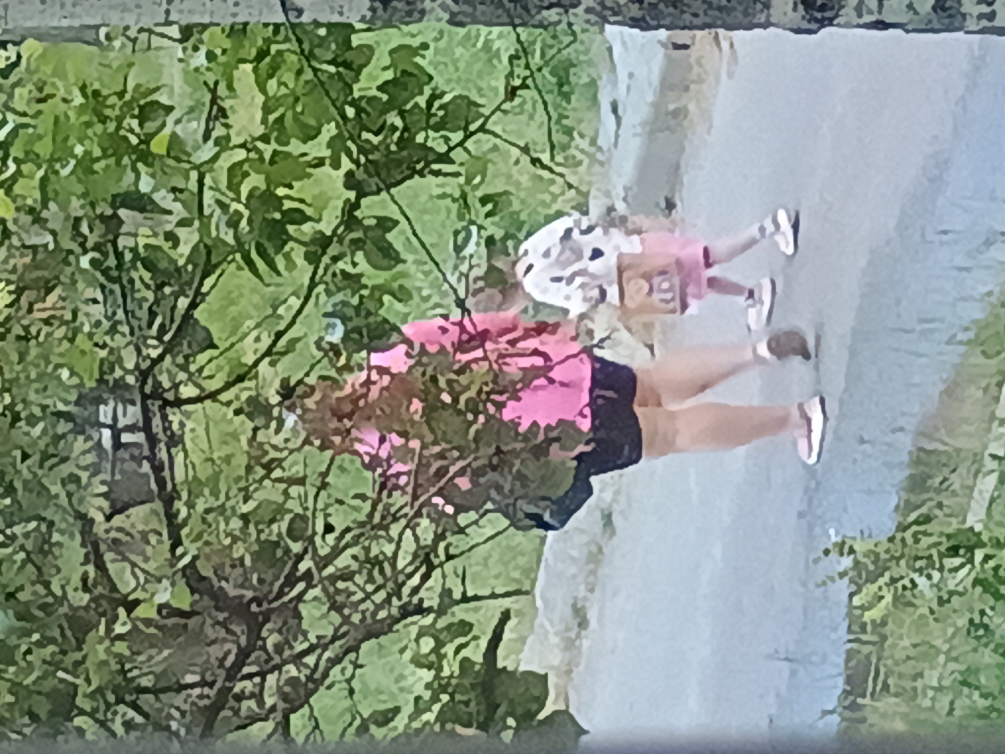 In an outdoor scene, possibly in Zaporizhzhia, Ukraine, two individuals are walking on a paved surface. The person in the foreground, on the left, is an adult wearing a bright pink long-sleeved shirt and dark shorts. They have a dark bag slung over their left shoulder. To their right, a child is walking, wearing a white shirt with a black heart pattern and pink shorts. The child is carrying a tan-colored bag. Both individuals are wearing athletic shoes. The background consists of green foliage from trees and shrubs, with a hint of a stone wall or embankment on the left. The lighting suggests it is daytime.