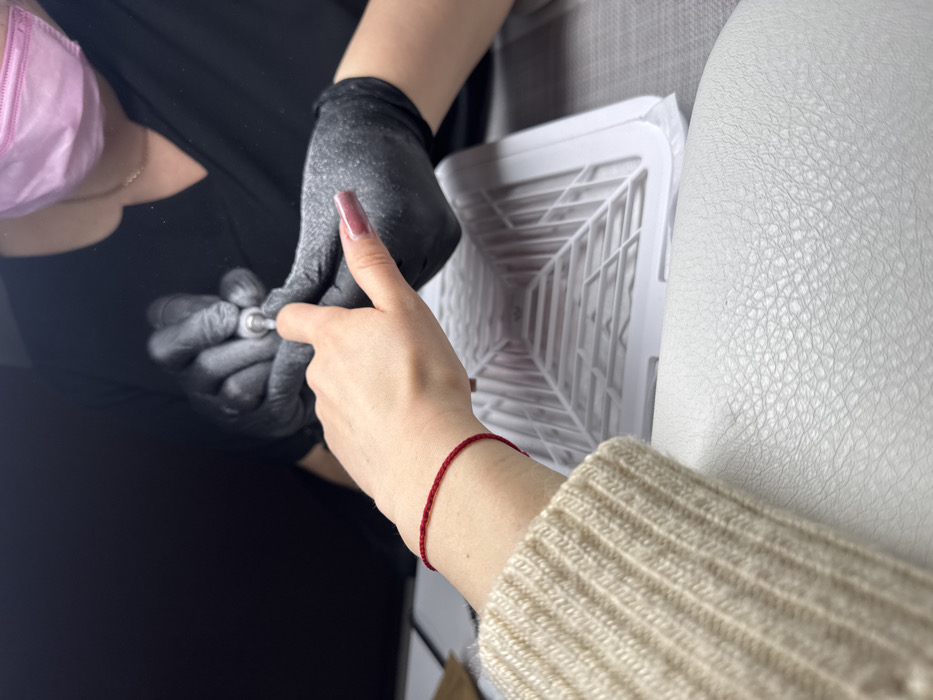 The scene depicts a nail care service taking place in a salon environment within the city of Unknown, Unknown. In the foreground, the left hand and forearm of a client are visible. The client wears a light beige ribbed knit sweater and a thin red string bracelet on the wrist. The client's left hand has natural or unfinished nails, with the exception of the thumb, which features a long, square-shaped nail painted with a glossy dark pink/mauve polish. This hand rests on a white, textured armrest or cushion. In the background, a nail technician, wearing a black short-sleeved top, black disposable gloves, and a light pink surgical mask, is actively performing a nail treatment. The technician's gloved hands manipulate an electric nail file on the client's left index finger, with fine dust particles visible around the rotating bit. A white, square-shaped nail dust collector with a gridded surface is situated on a dark tabletop between the client's hand and the technician's workspace. The background is dark and out of focus.